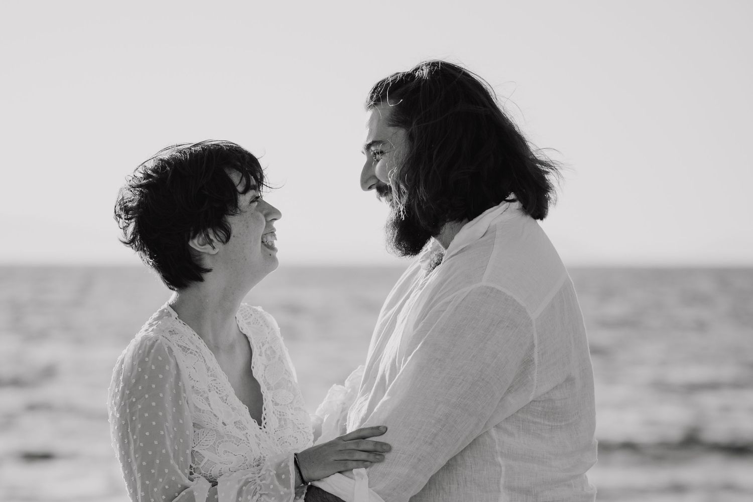 Black and white photo of a couple embracing and smiling at each other on a beach by the ocean.