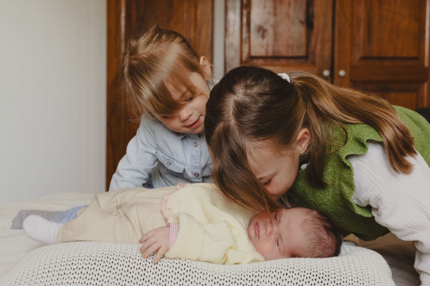 Two young children lovingly leaning over a sleeping newborn baby on a white blanket.
