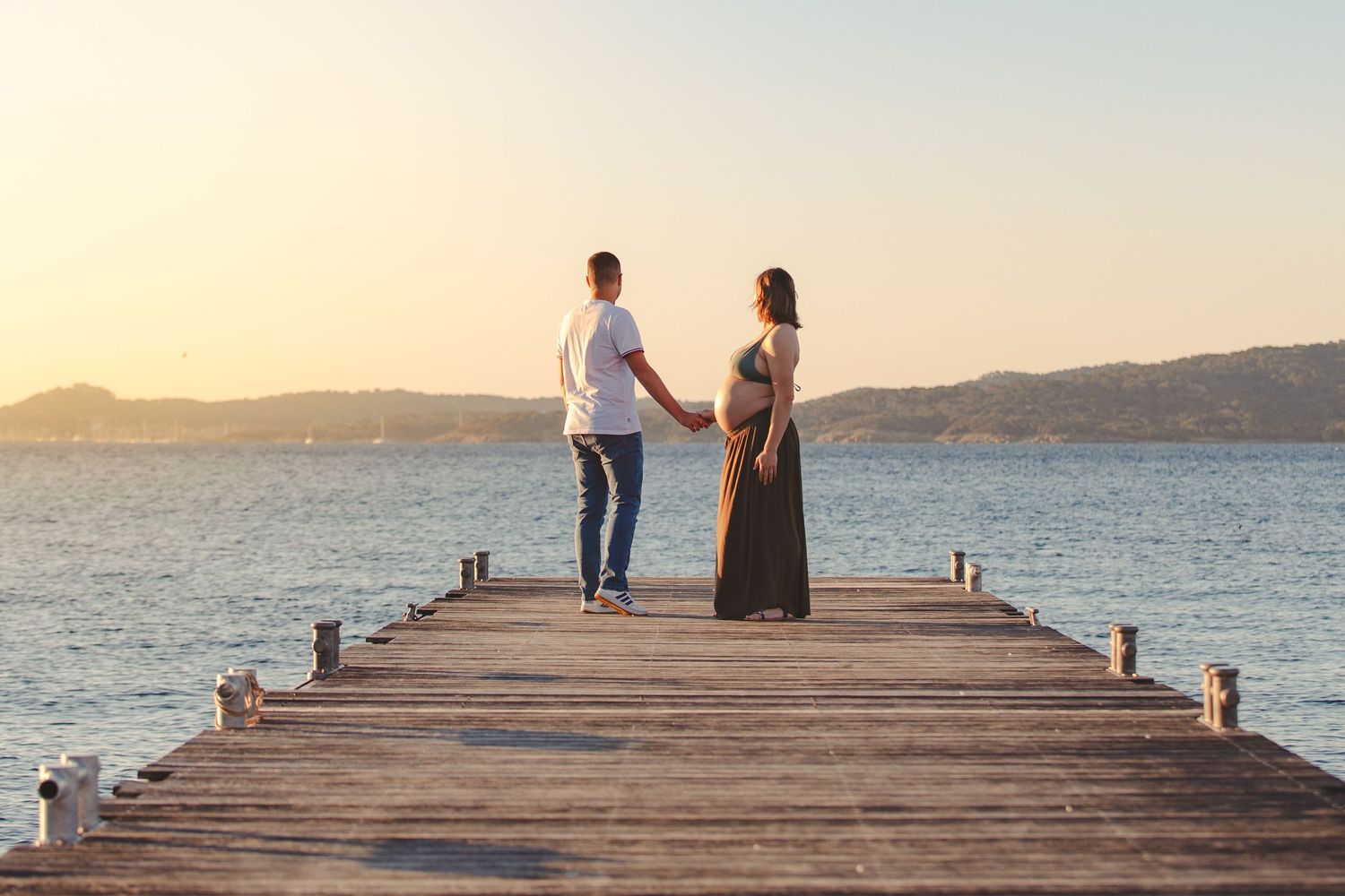 Couple holding hands on wooden pier at sunset, pregnant woman in dark dress facing calm water and distant hills.