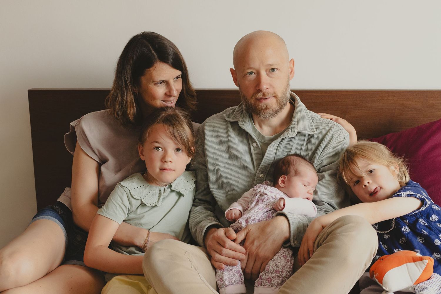 Family of five with parents and three children including a newborn sitting together on a bed.
