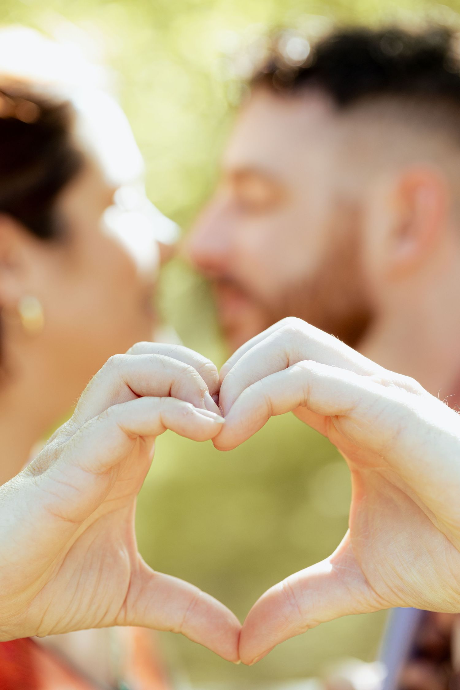 Couple kissing while making a heart shape with their hands outdoors in a sunny park.
