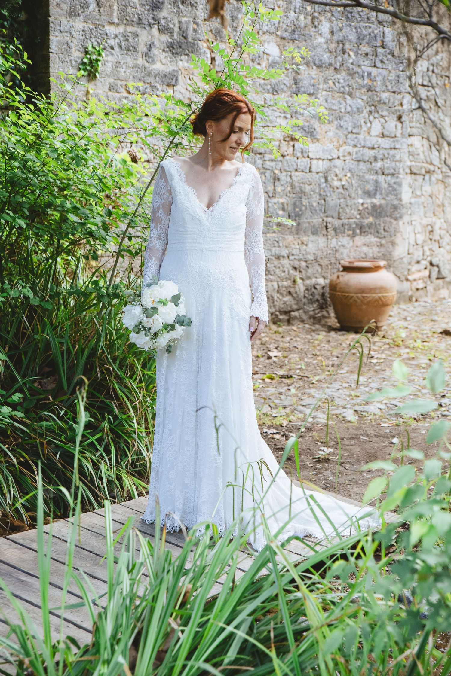 Bride in white lace long-sleeve gown holding bouquet stands on wooden path in lush garden near stone wall.