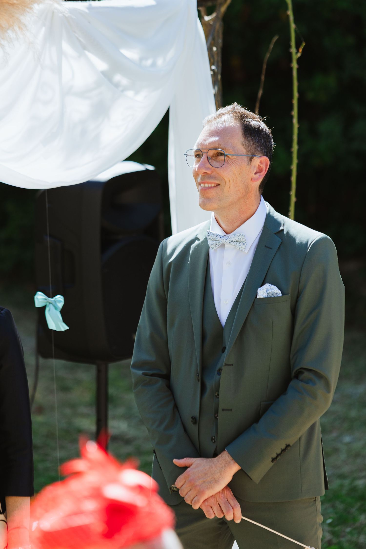 Smiling groom in green suit with bow tie standing at outdoor wedding ceremony.