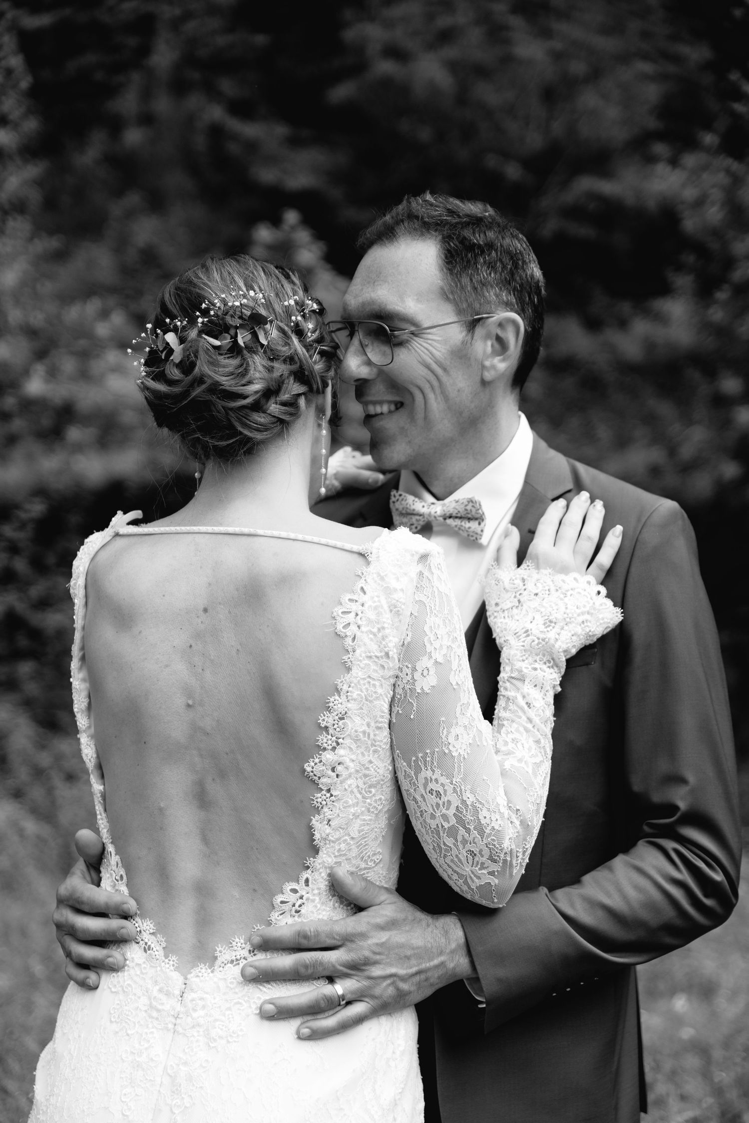 Black and white wedding photo of bride in lace gown and groom in suit sharing a romantic embrace outdoors.