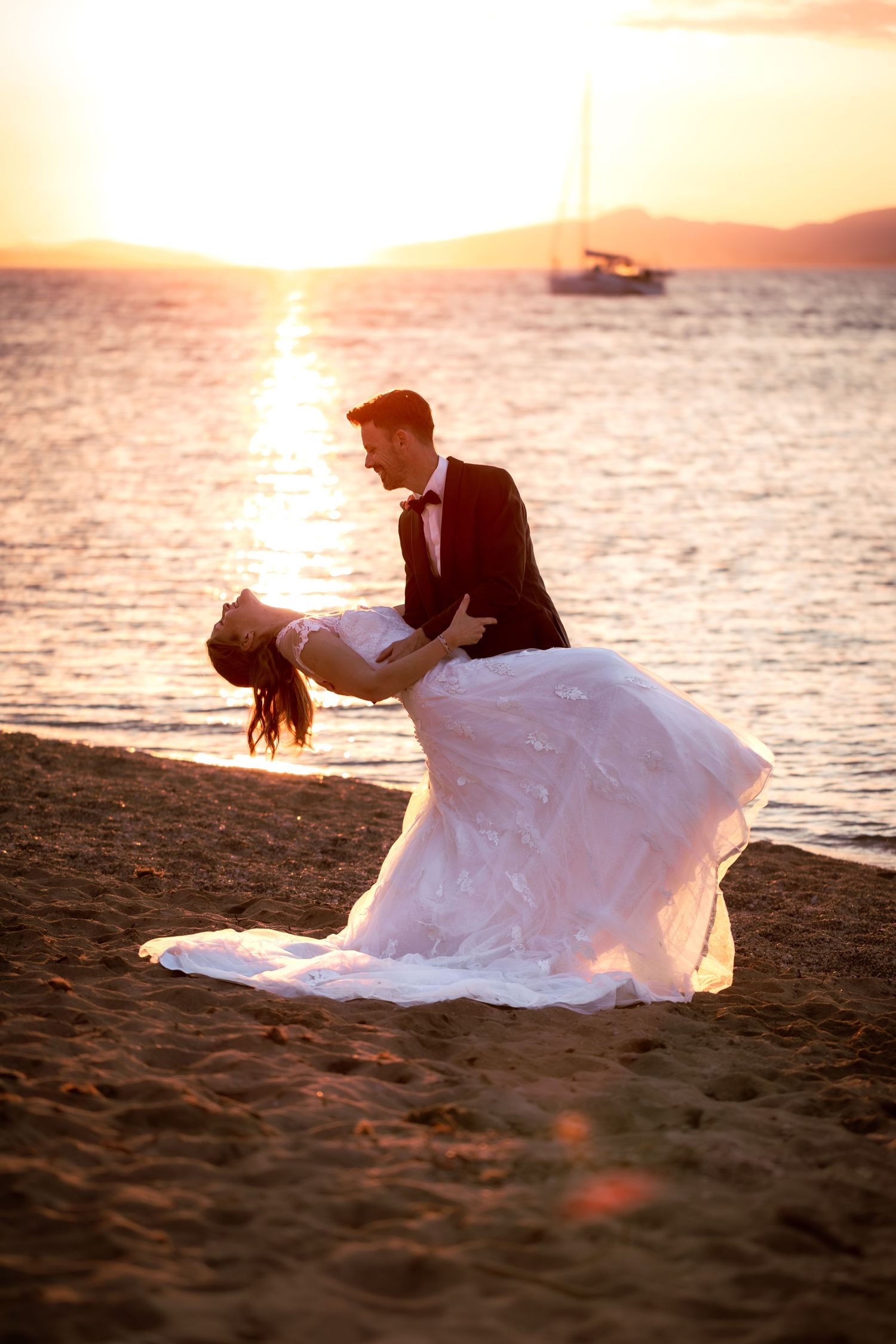 Bride and groom dancing on a sandy beach at golden sunset, with sailboat and mountains in background.