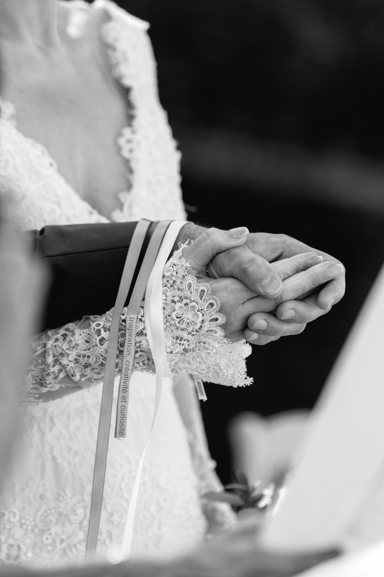 Black and white photo of bride and groom holding hands during wedding ceremony, bride wearing lace dress.