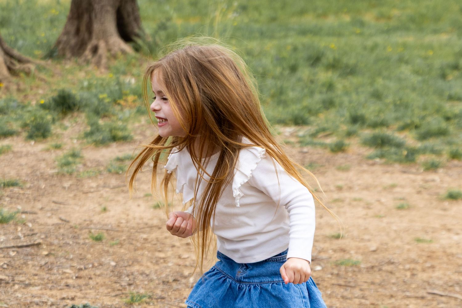 Young girl with long hair runs and laughs outdoors in a park wearing a denim skirt.