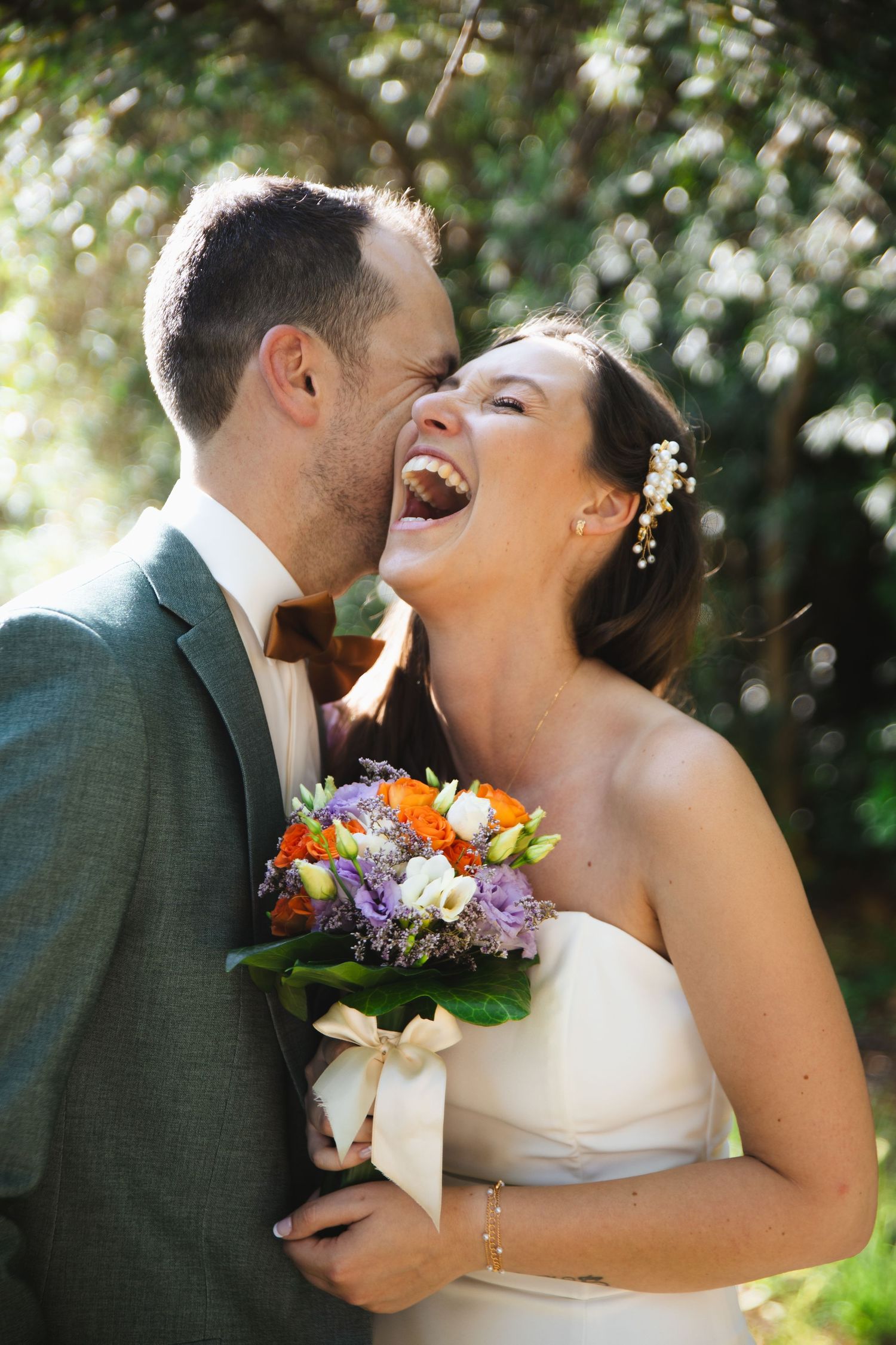 Groom whispering in brides ear as she laughs holding colorful bouquet in sunny garden setting.