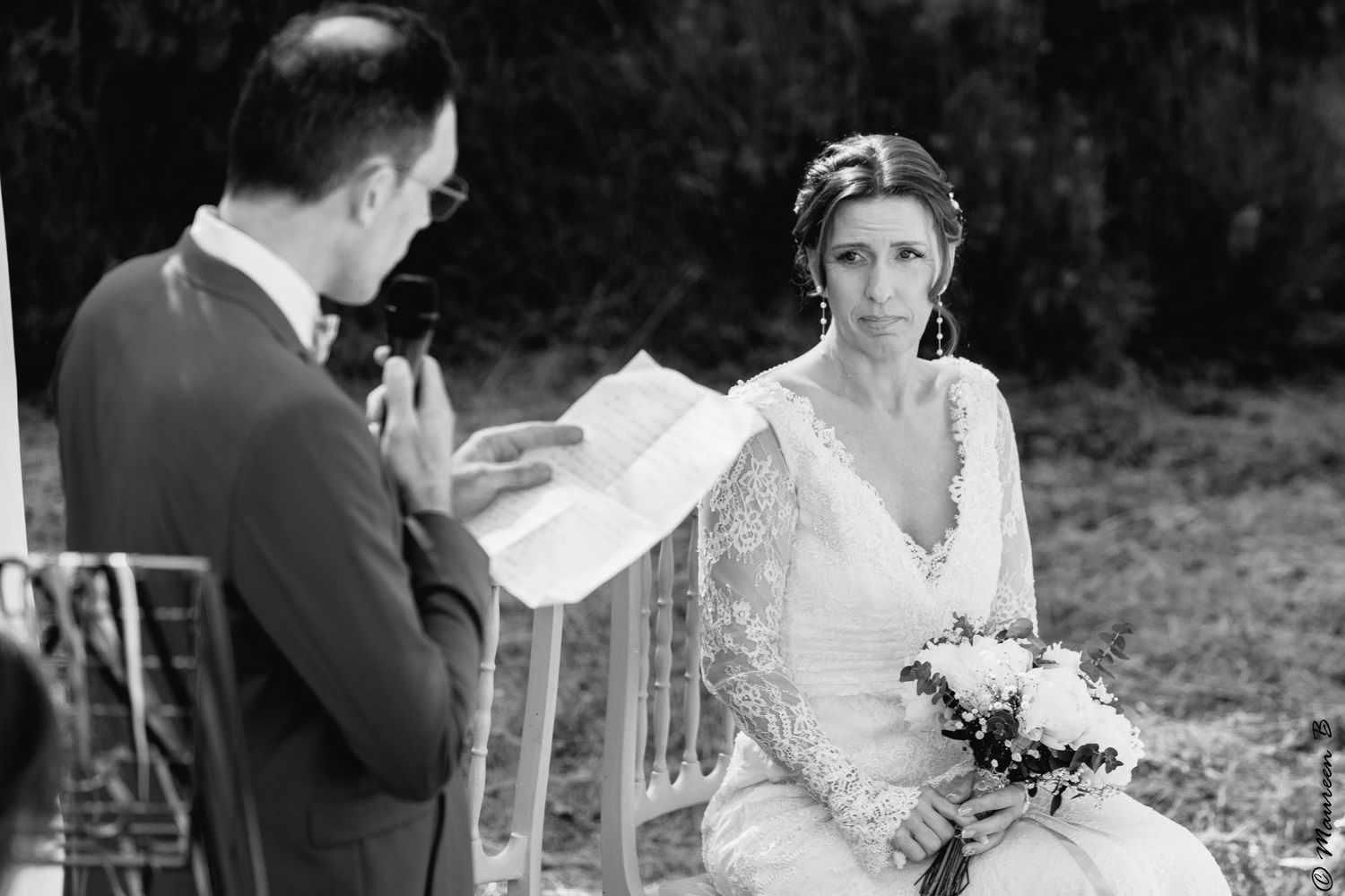 Bride smiles emotionally as groom reads wedding vows during an outdoor black and white ceremony photo.