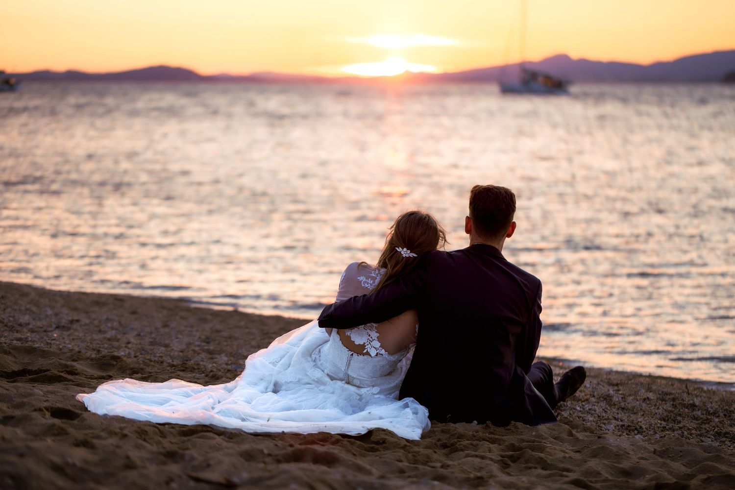 Couple in wedding attire sitting together on a sandy beach watching a romantic golden sunset over calm ocean waters.