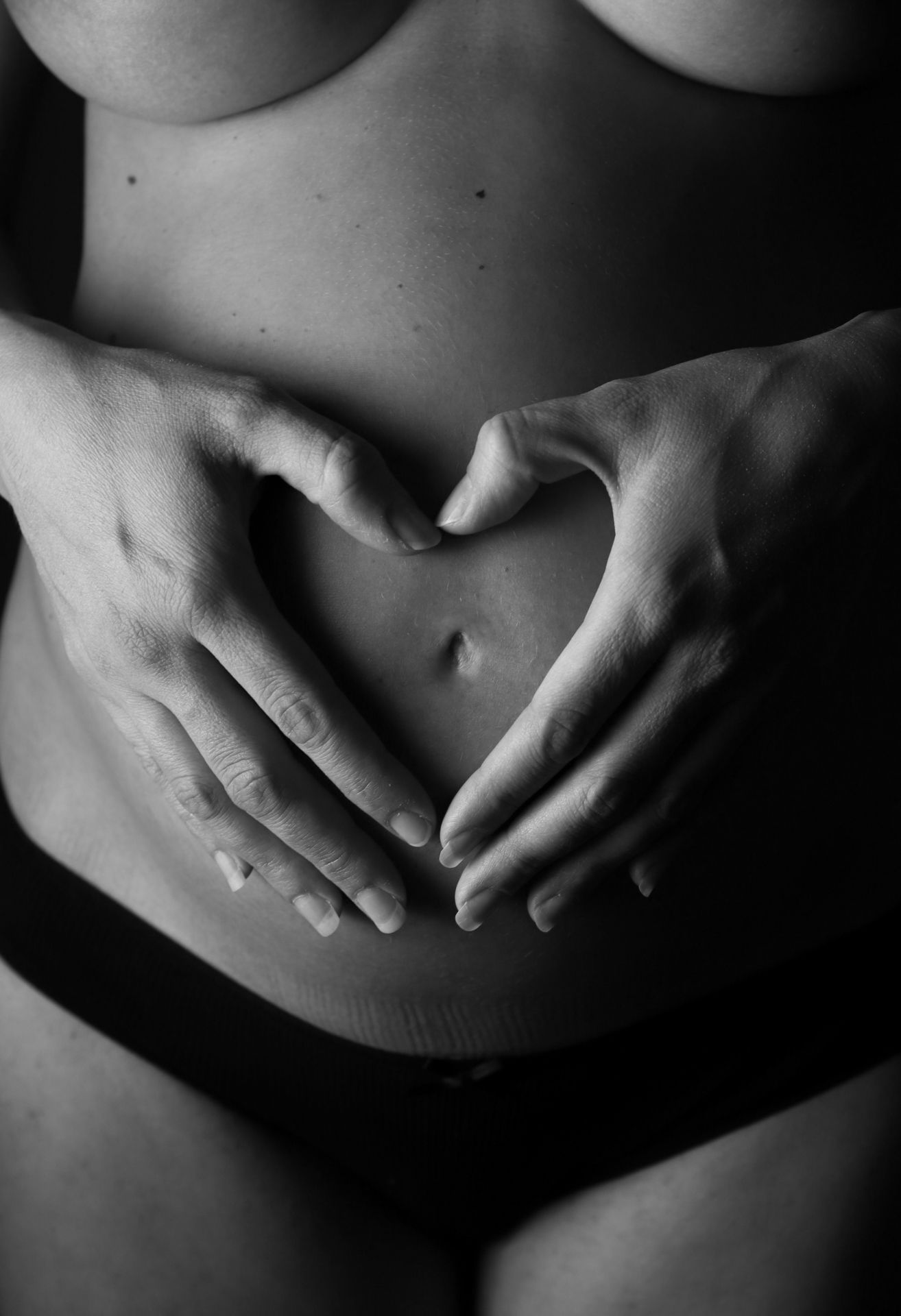 Close-up black and white photo of pregnant woman forming heart shape on belly.