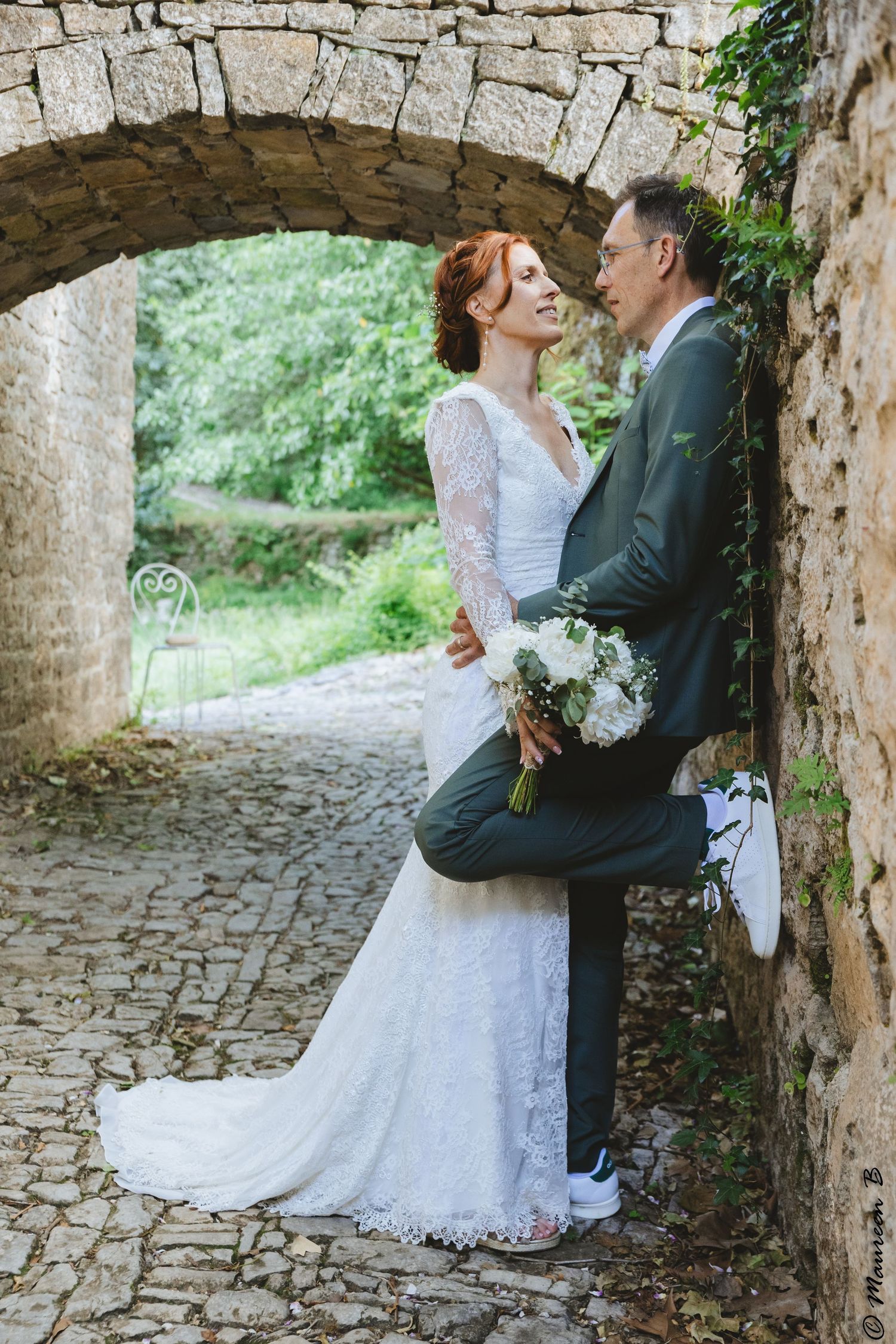 Bride and groom share a romantic moment under a stone arch on a cobblestone path outdoors.