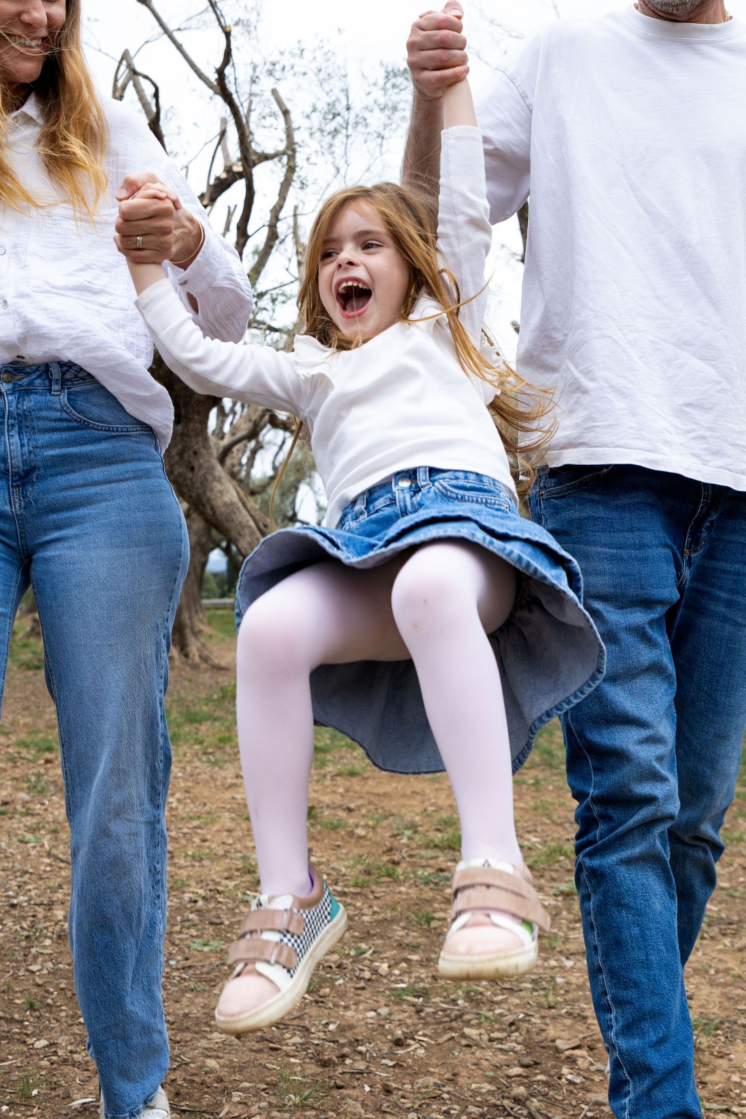 Parents swing their laughing daughter by the hands in an outdoor park setting.