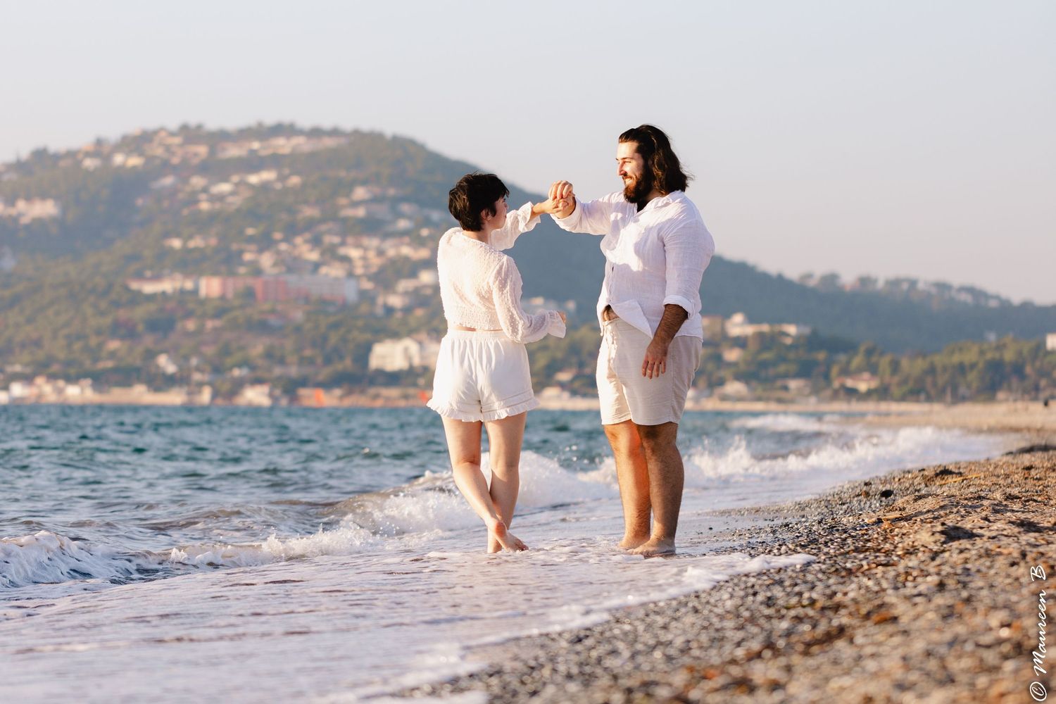 Happy couple in white outfits holding hands and dancing on a scenic pebble beach.
