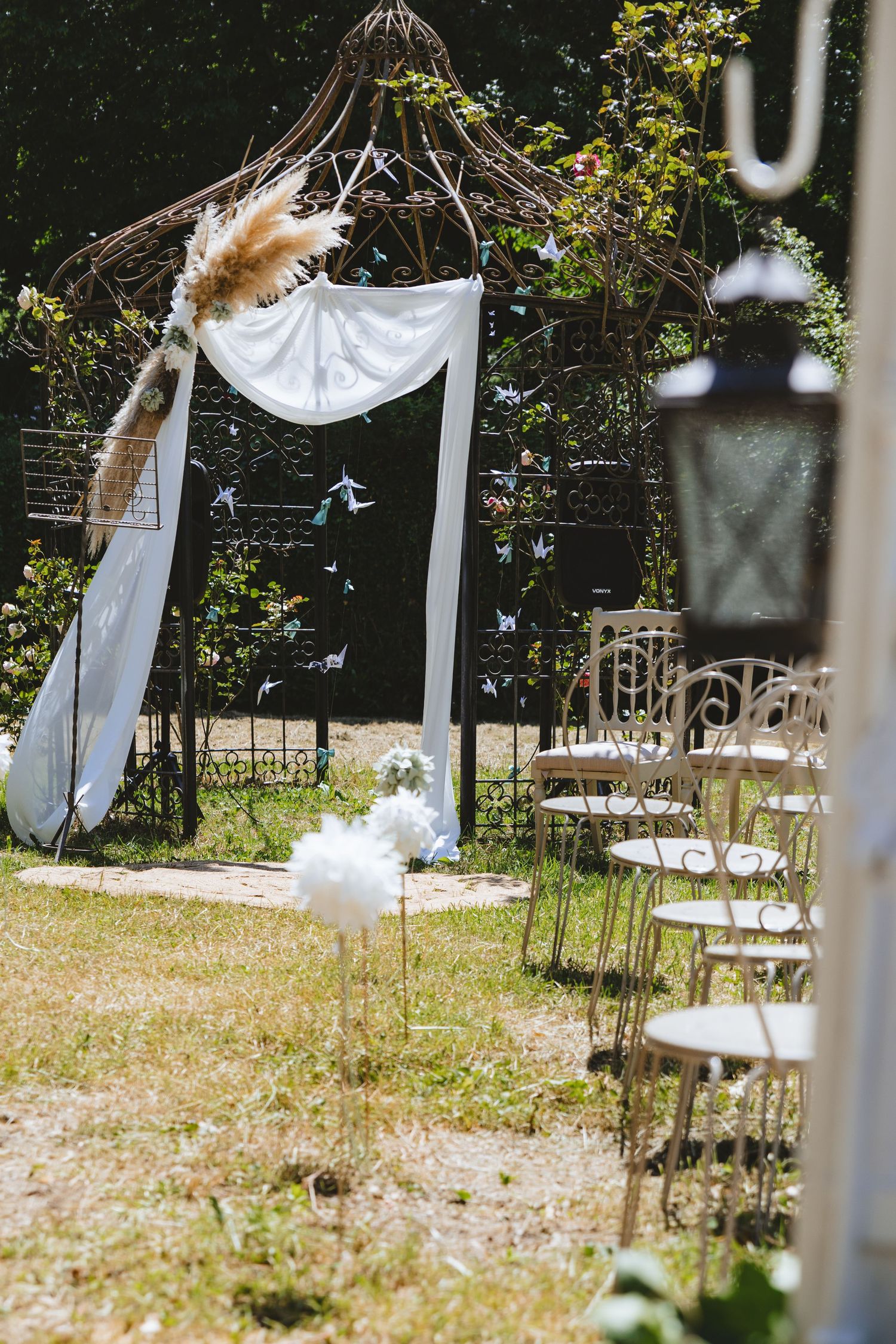Outdoor wedding ceremony arch decorated with white draping, pampas grass, and paper birds, with white chairs on a sunny lawn.
