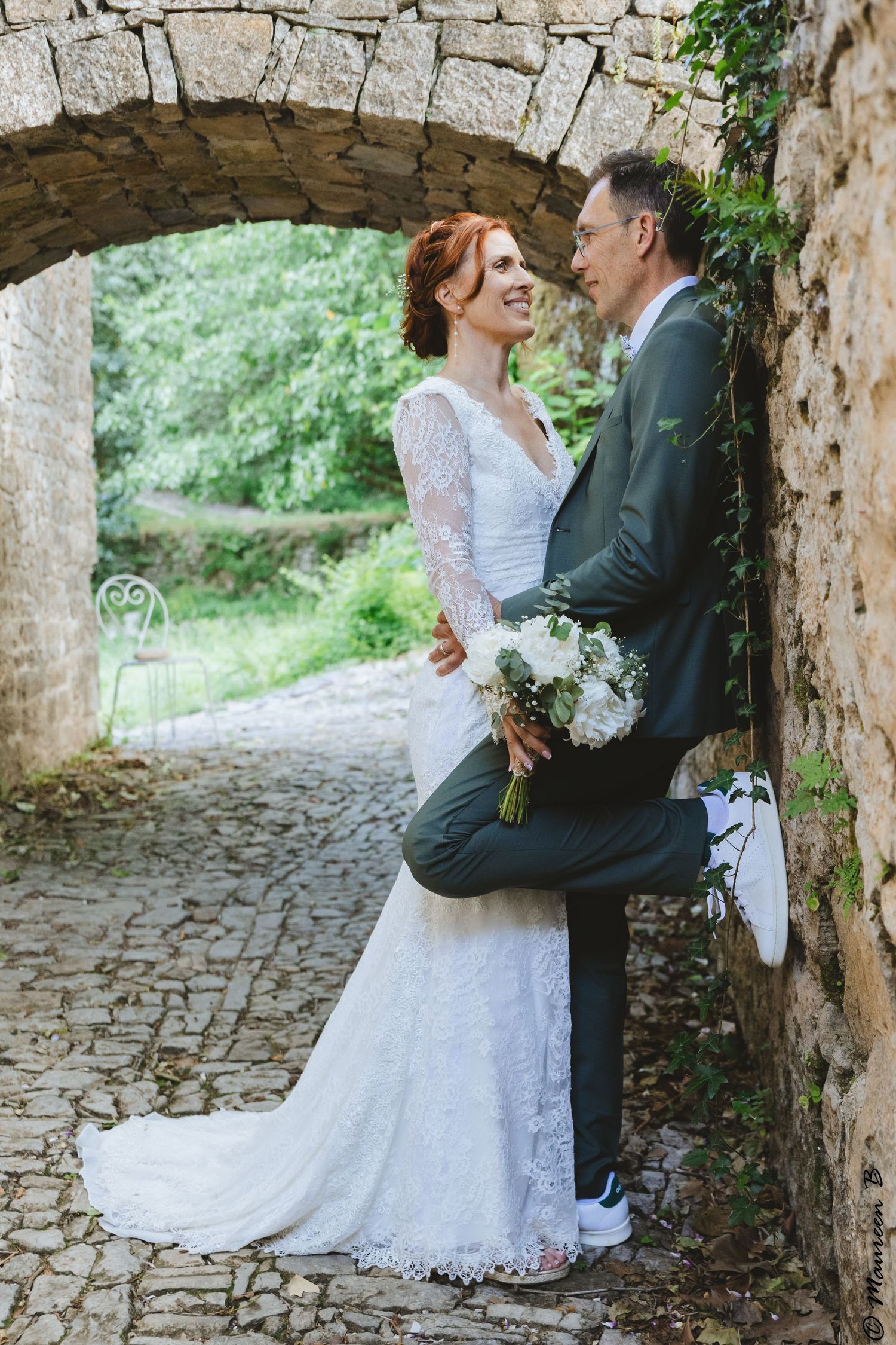 Bride in lace gown and groom in gray suit share a romantic moment under a stone arch with greenery.