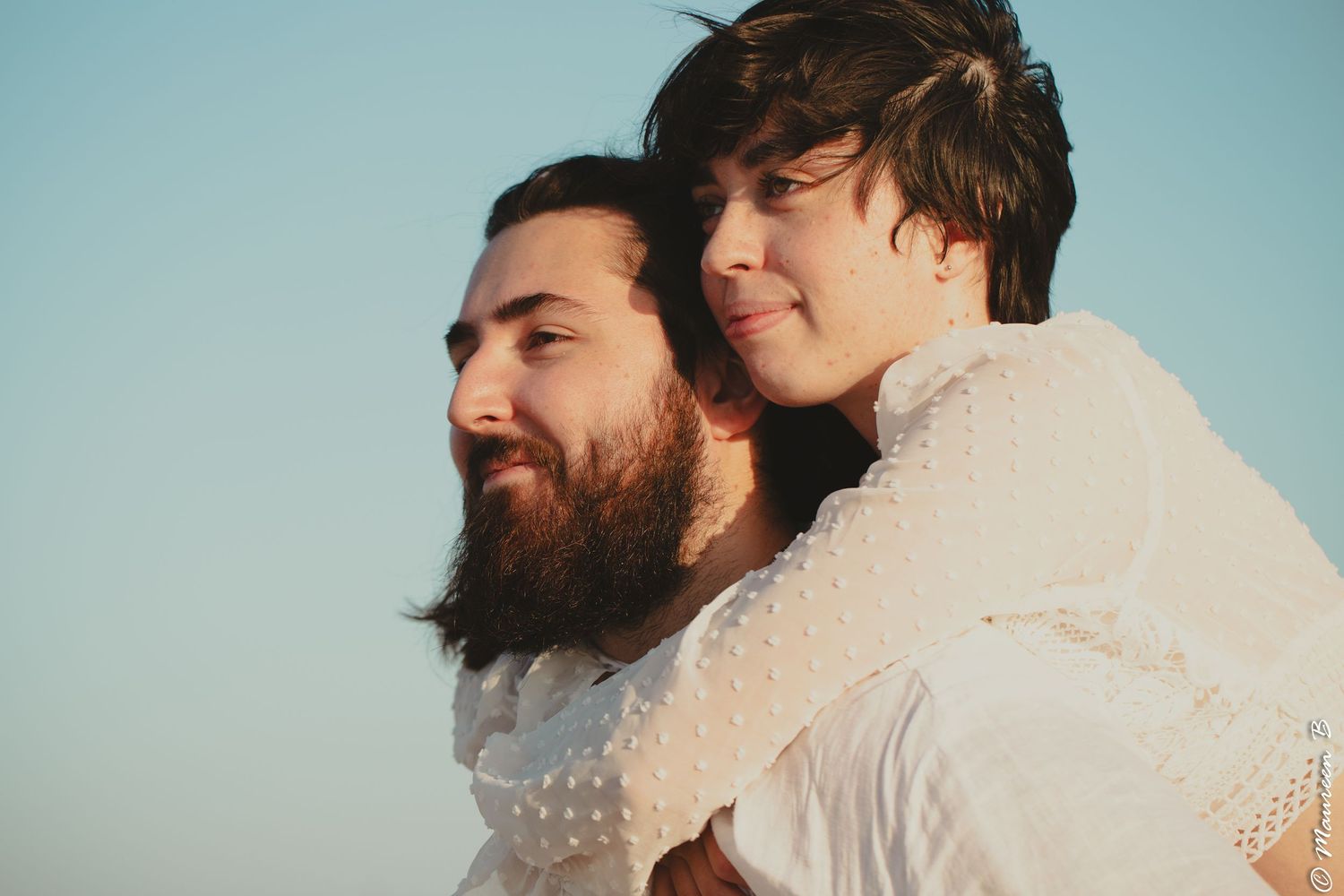 Bearded man giving a woman a piggyback ride against a clear blue sky outdoors.
