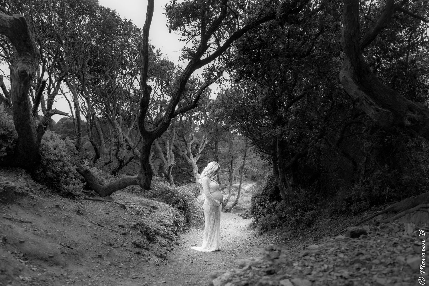 Black and white photo of a pregnant woman in a white dress standing on a forest path.