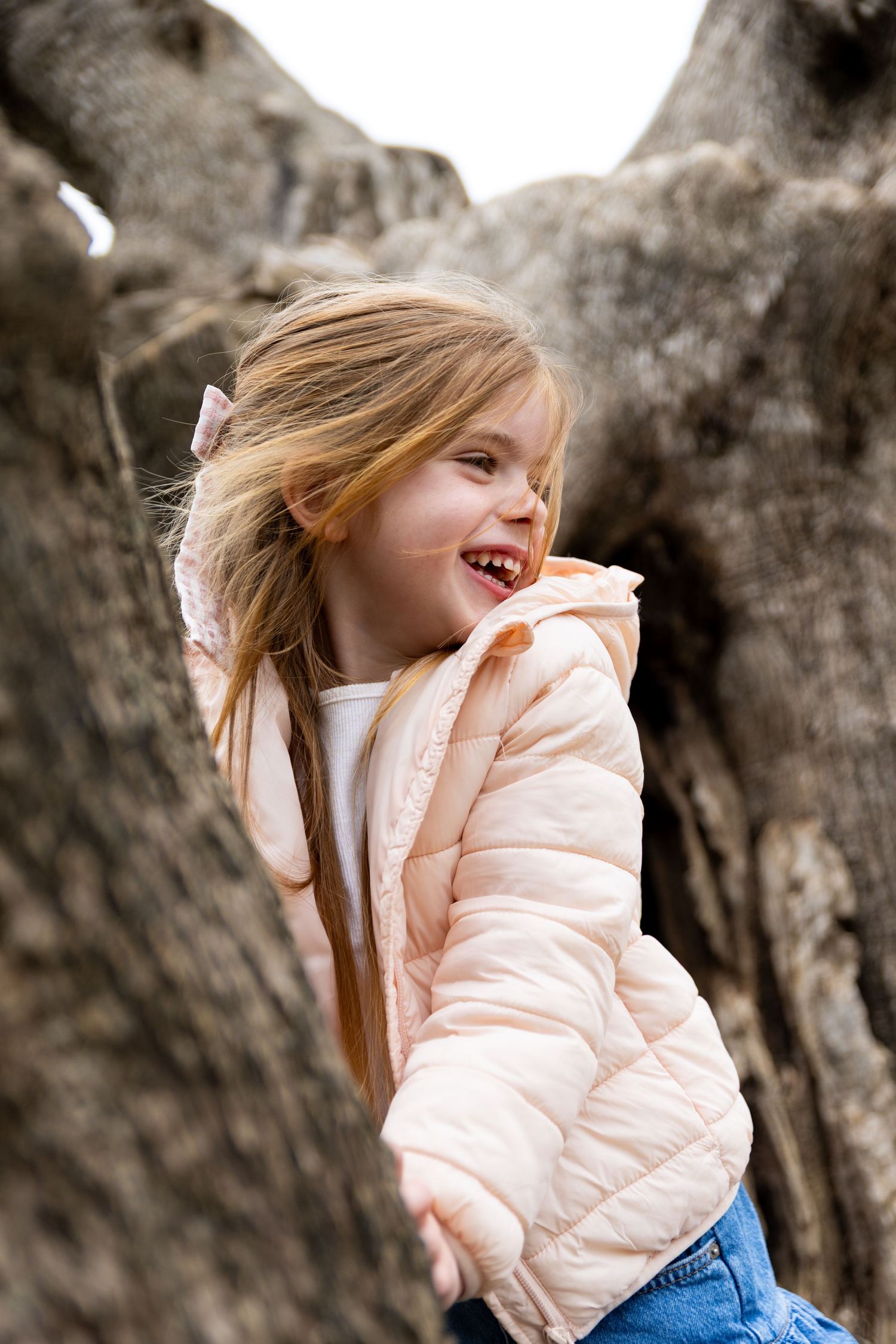A cheerful young girl in a pink jacket laughs joyfully while playing near a large tree trunk outdoors.