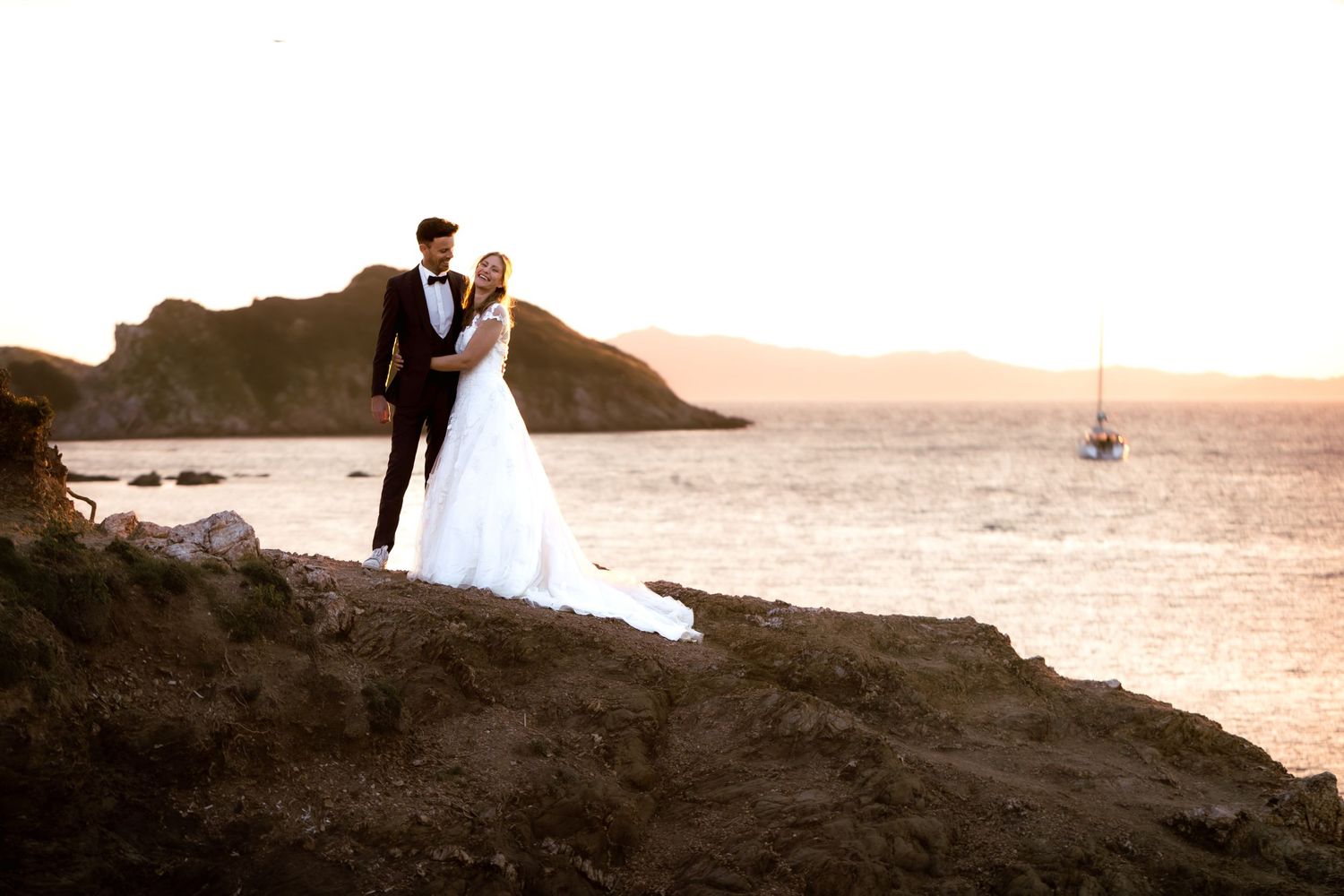 Bride and groom pose on rocky coastal cliffs at golden sunset with sailboat and mountains in background.