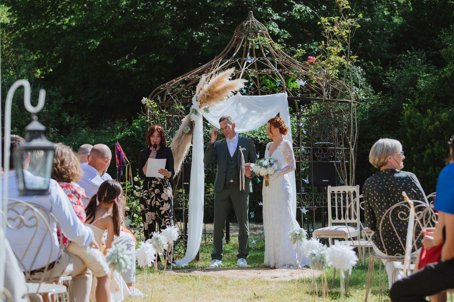 Outdoor wedding ceremony under rustic arch with pampas grass, bride and groom standing before officiant in sunny garden.