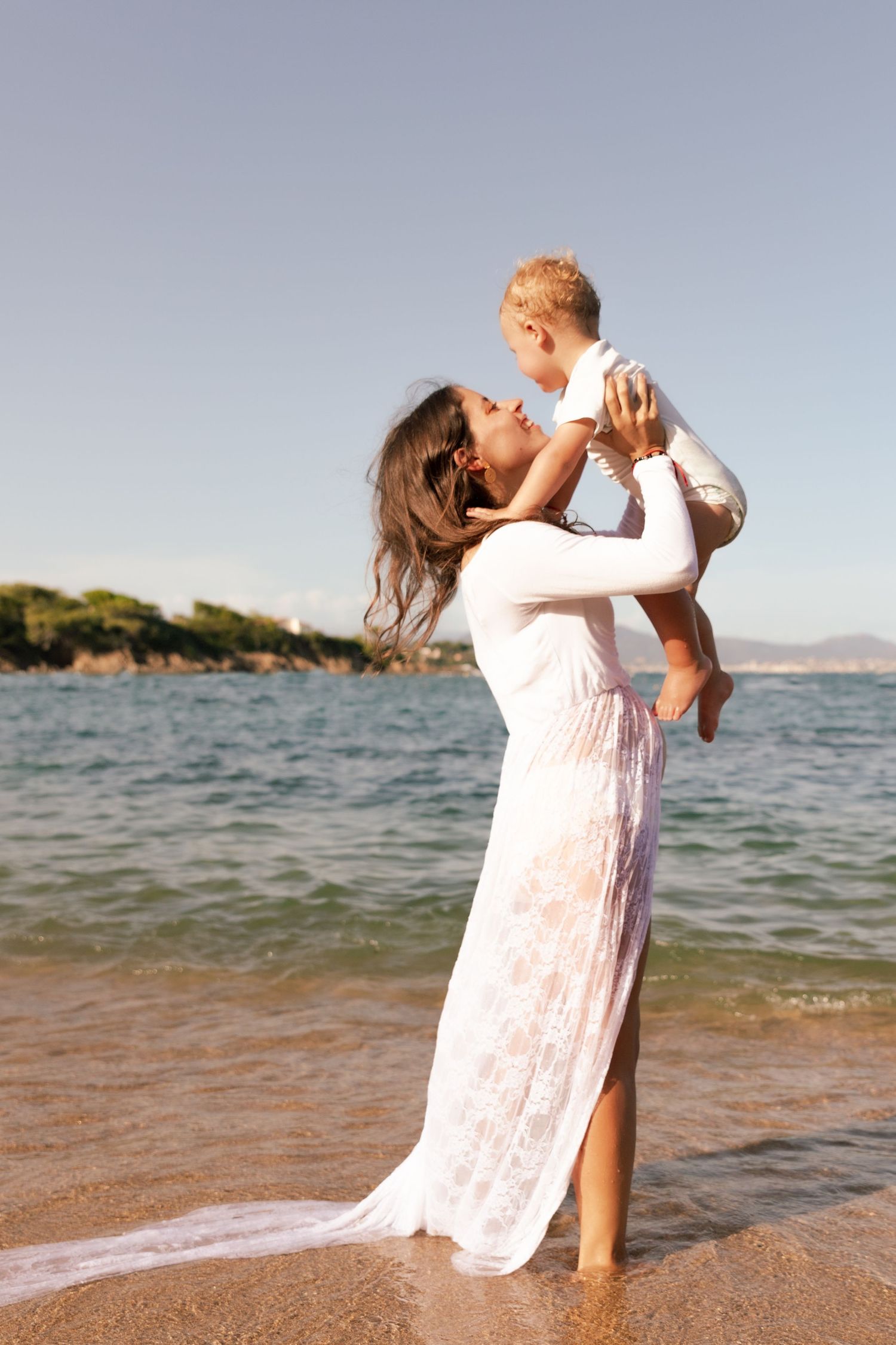 A mother in a white lace dress lifts her baby joyfully on a sunny beach with blue ocean water behind them.