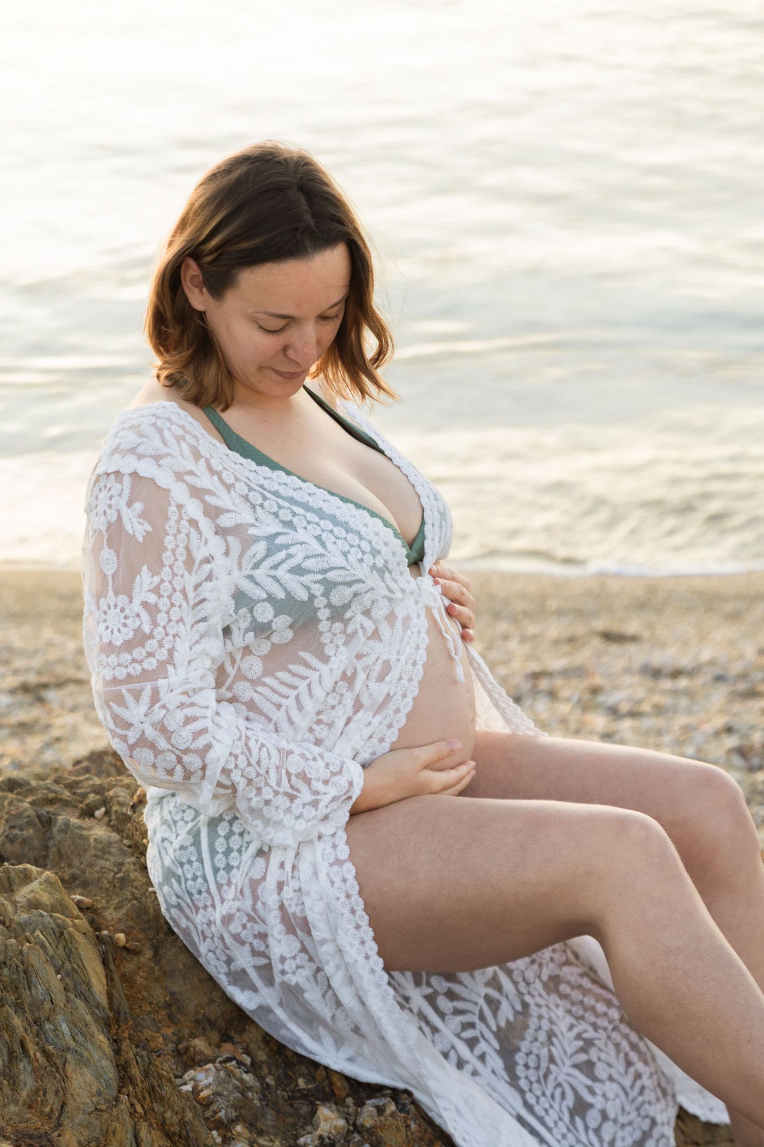 Pregnant woman in white lace cover-up sitting on beach rock, gently cradling her belly by the ocean.