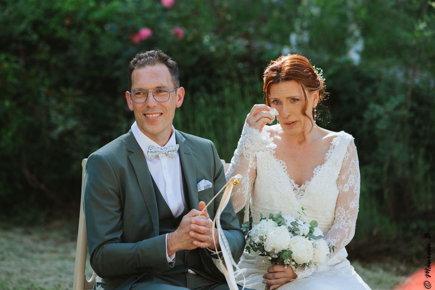 Bride wipes tears while groom smiles holding champagne during an outdoor wedding ceremony in a garden.