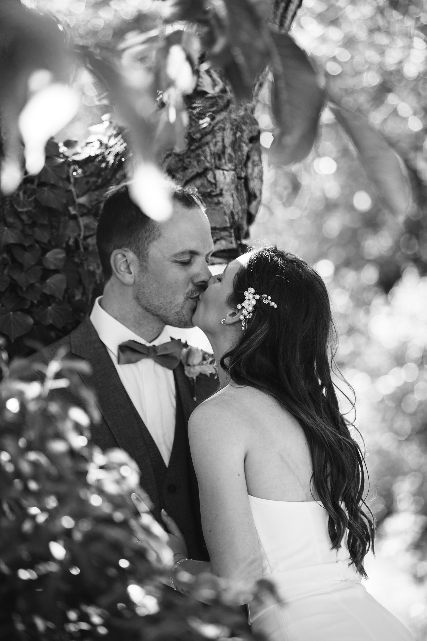 Black and white photo of bride and groom sharing a kiss beneath lush tree branches.
