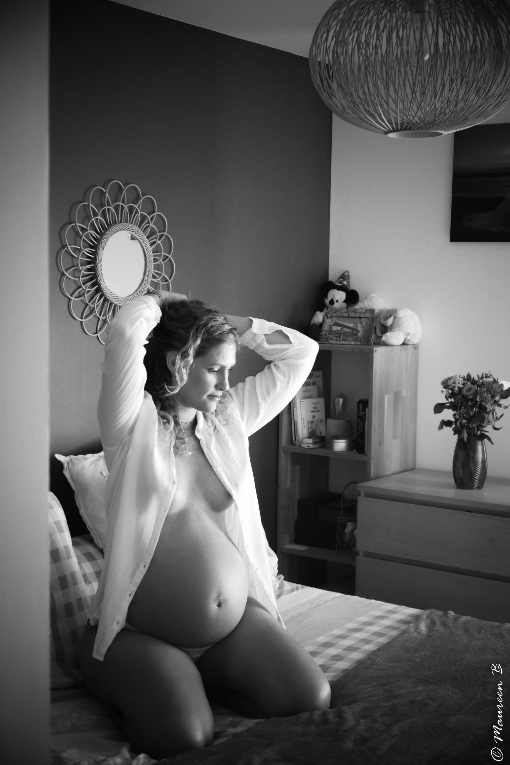 Black and white maternity photo of pregnant woman kneeling on bed in open white shirt.