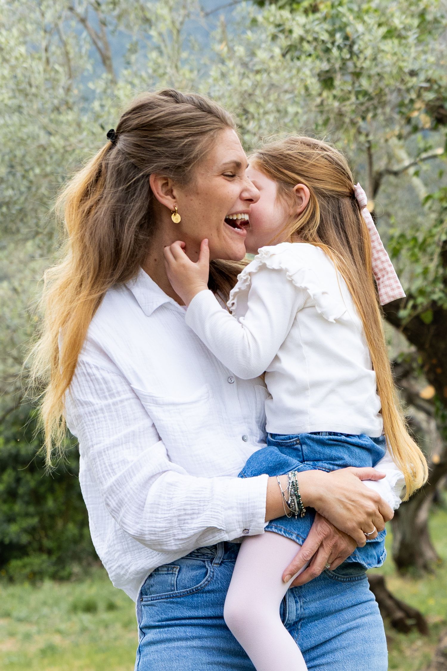 Mother holds young daughter who playfully licks her cheek outdoors in an olive grove.