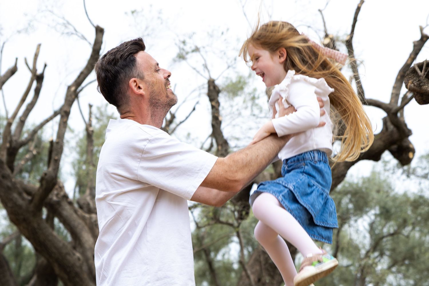 Father lifts laughing young girl into the air outdoors among trees on a sunny day.