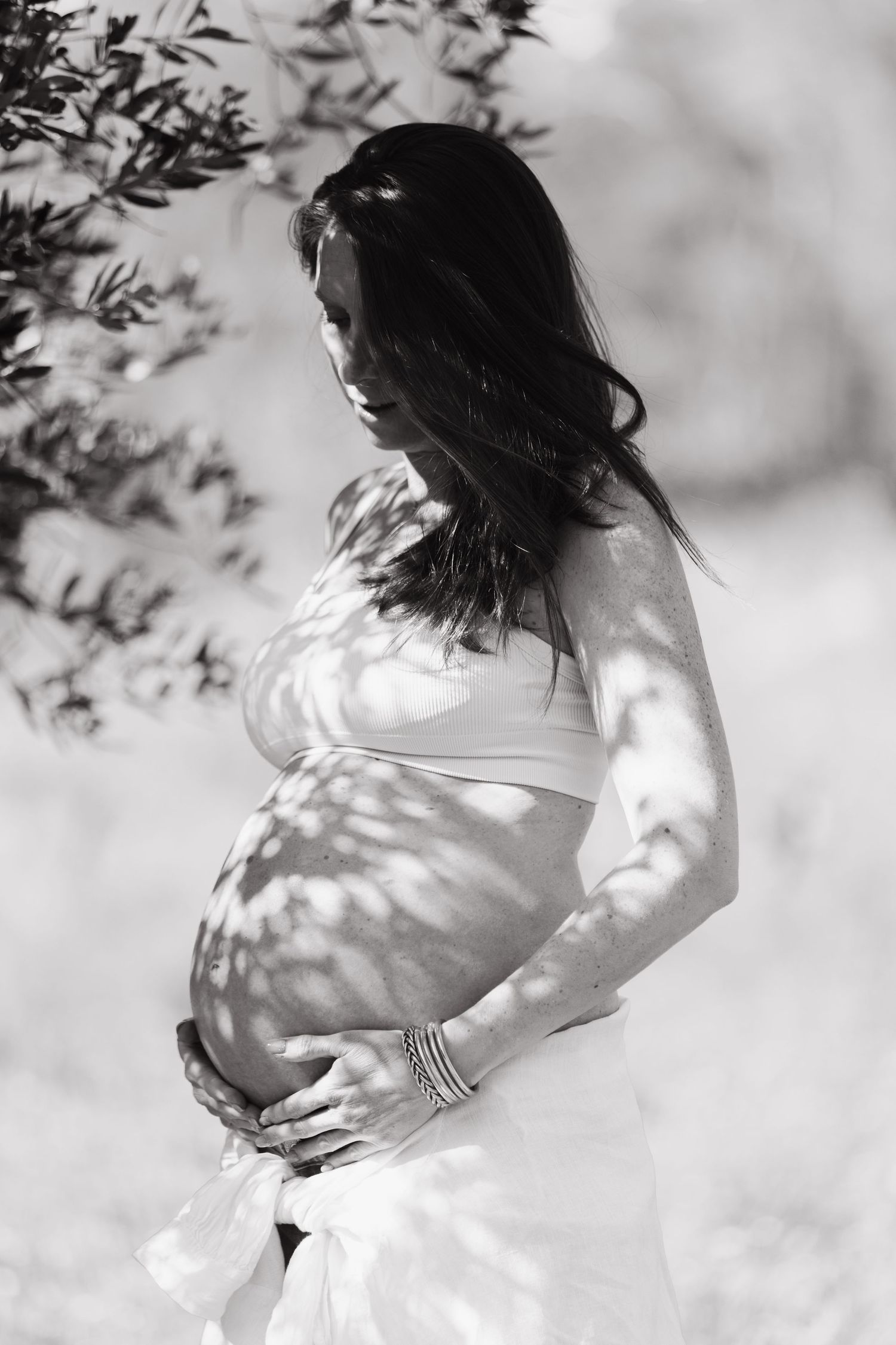 Black and white portrait of pregnant woman cradling baby bump under dappled tree light.