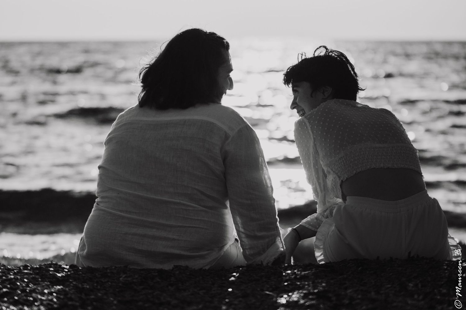 Black and white photo of a couple sitting together on a pebble beach facing the sea.
