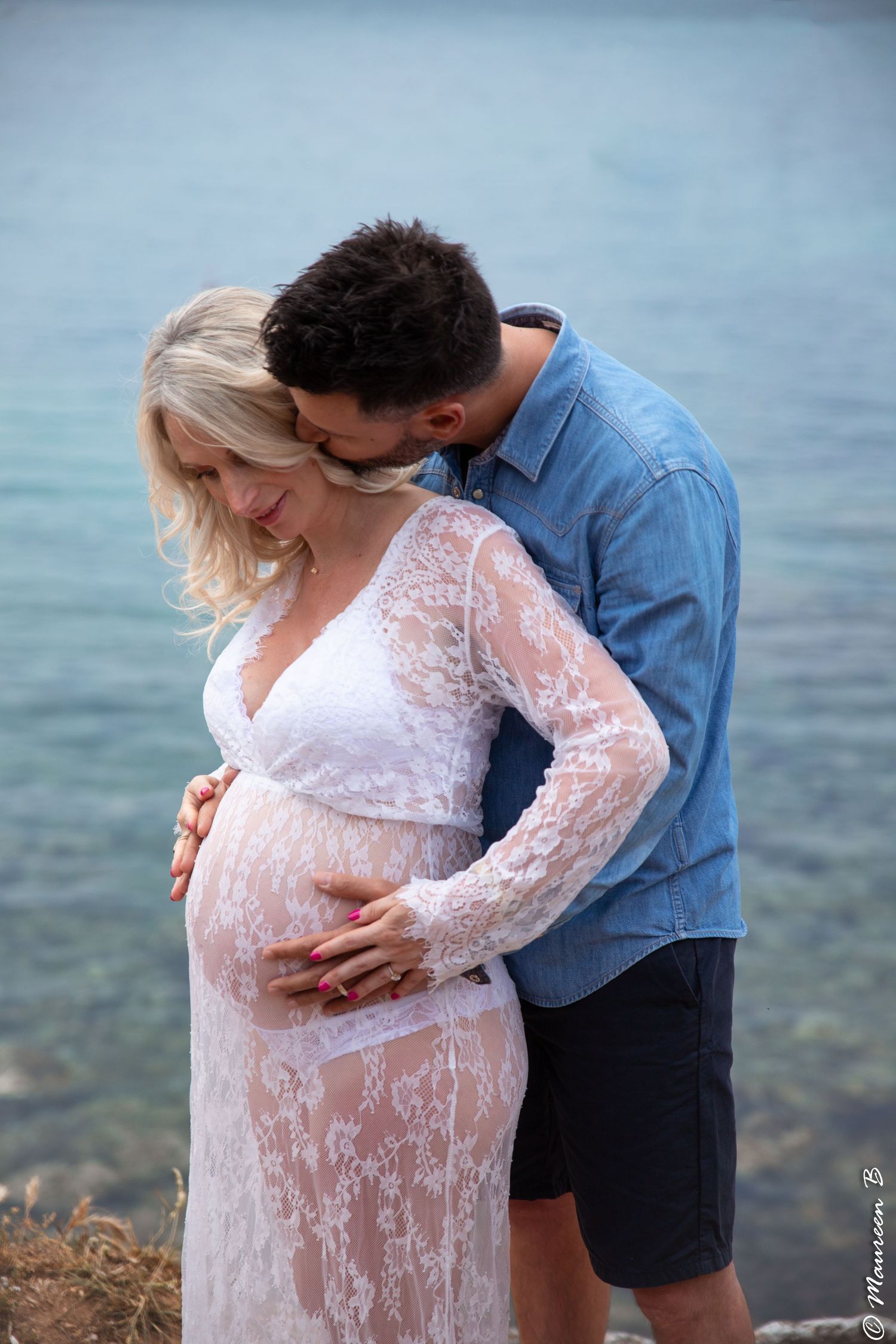 Couple sharing tender maternity photo by the sea, man kissing pregnant woman in white lace dress.
