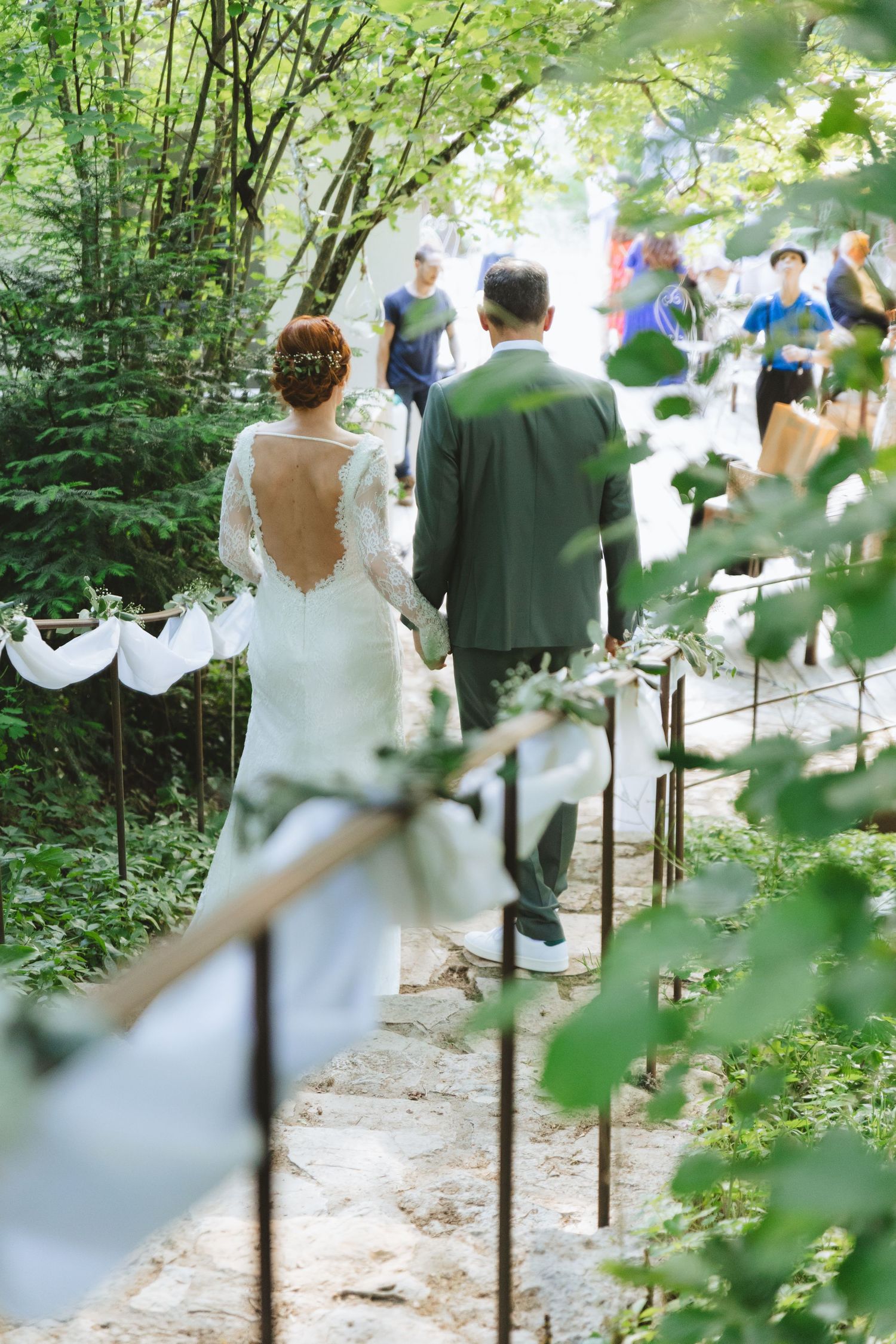 Bride and groom walk hand-in-hand down a stone path surrounded by lush green trees at an outdoor wedding ceremony.