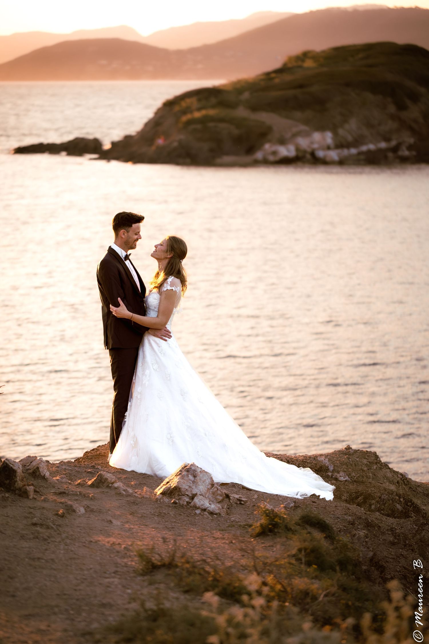 Bride and groom embrace on rocky coastline at golden sunset with island backdrop.