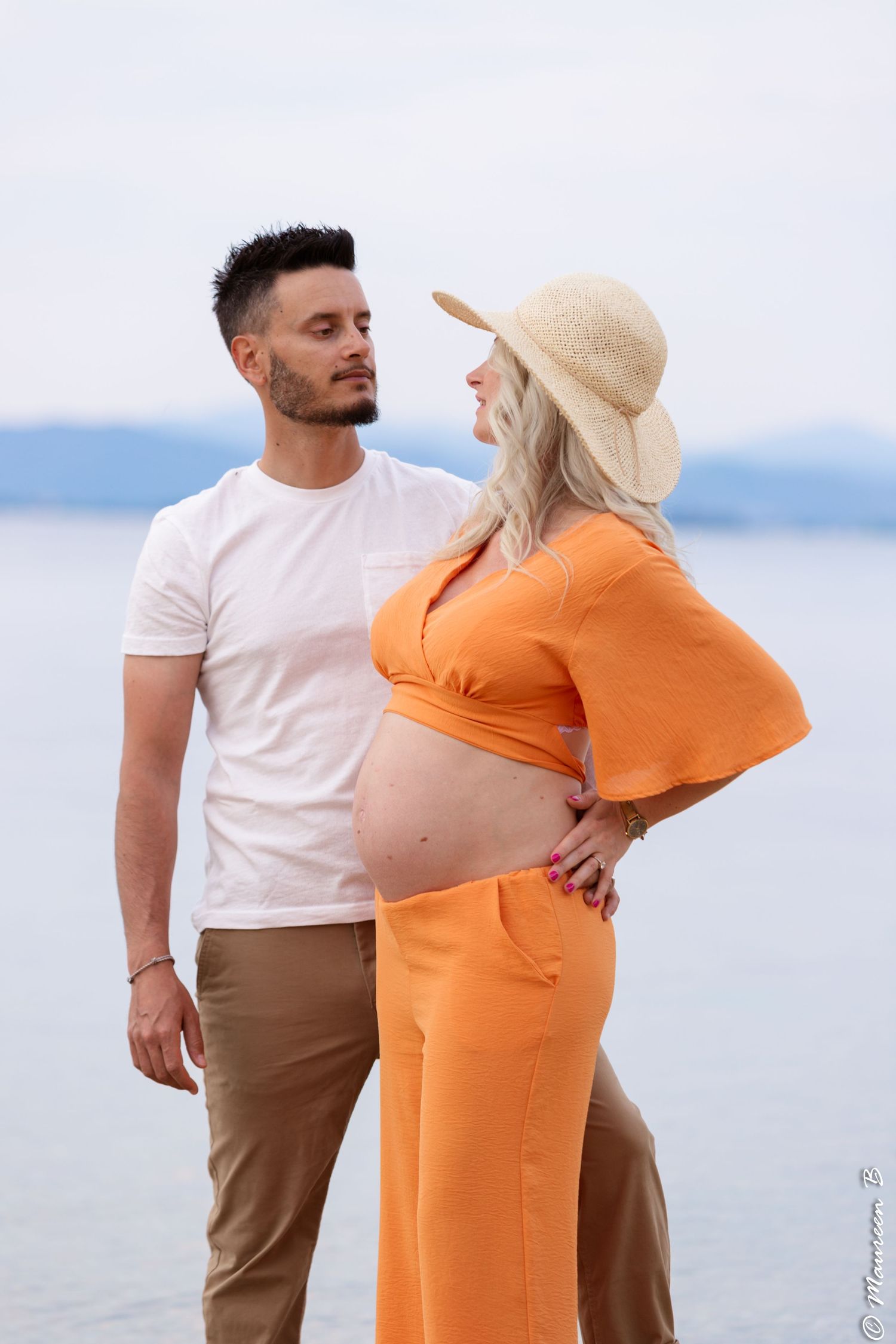 Couple sharing a loving glance outdoors by the sea, pregnant woman in orange outfit and straw hat.