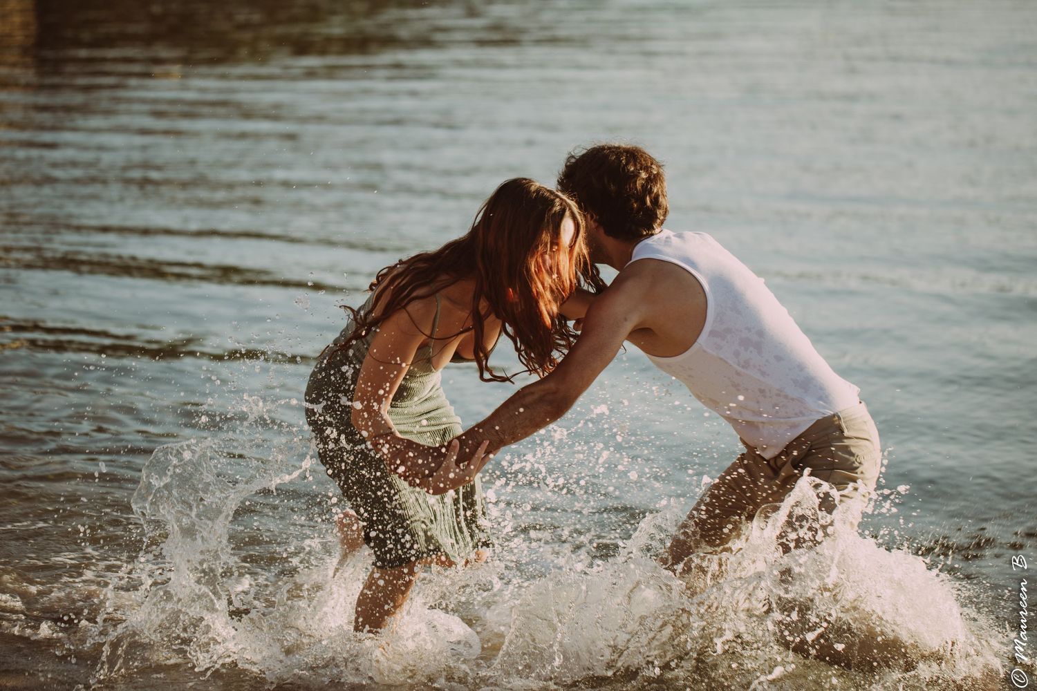 Playful couple splashing and laughing together in sparkling sunlit lake waters on a warm day.