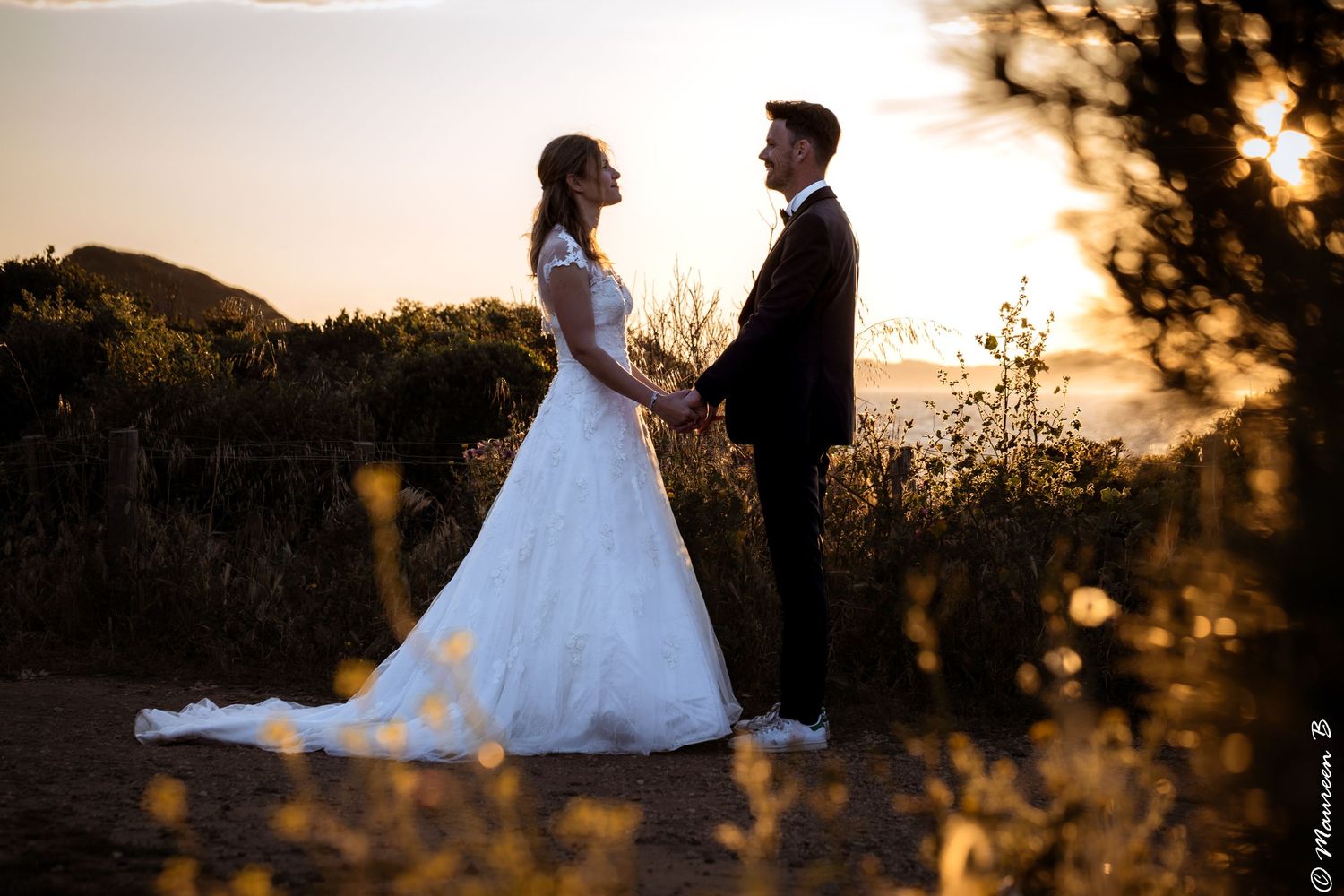 Bride and groom facing each other in golden sunset light among wildflowers in romantic portrait.