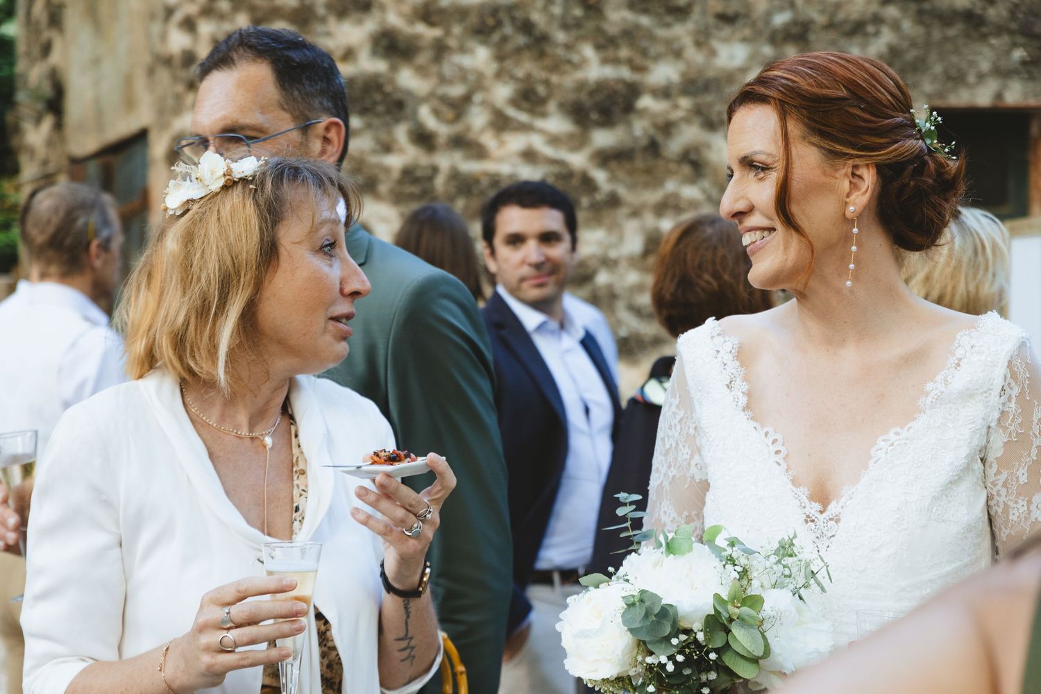 Bride in lace gown holds white bouquet, smiling with guests at an outdoor wedding reception.
