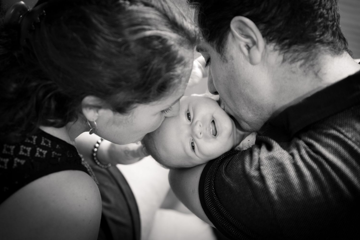 Black and white photo of two parents lovingly kissing their smiling baby on the cheeks simultaneously.