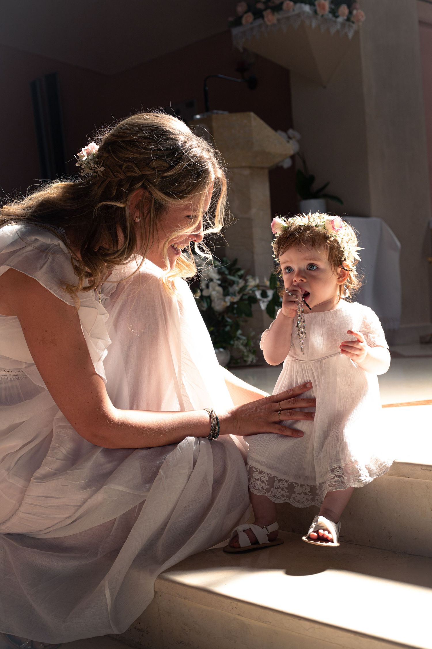 A smiling woman in white dress crouches beside a toddler girl in white lace dress, sharing a tender moment in sunlit church.