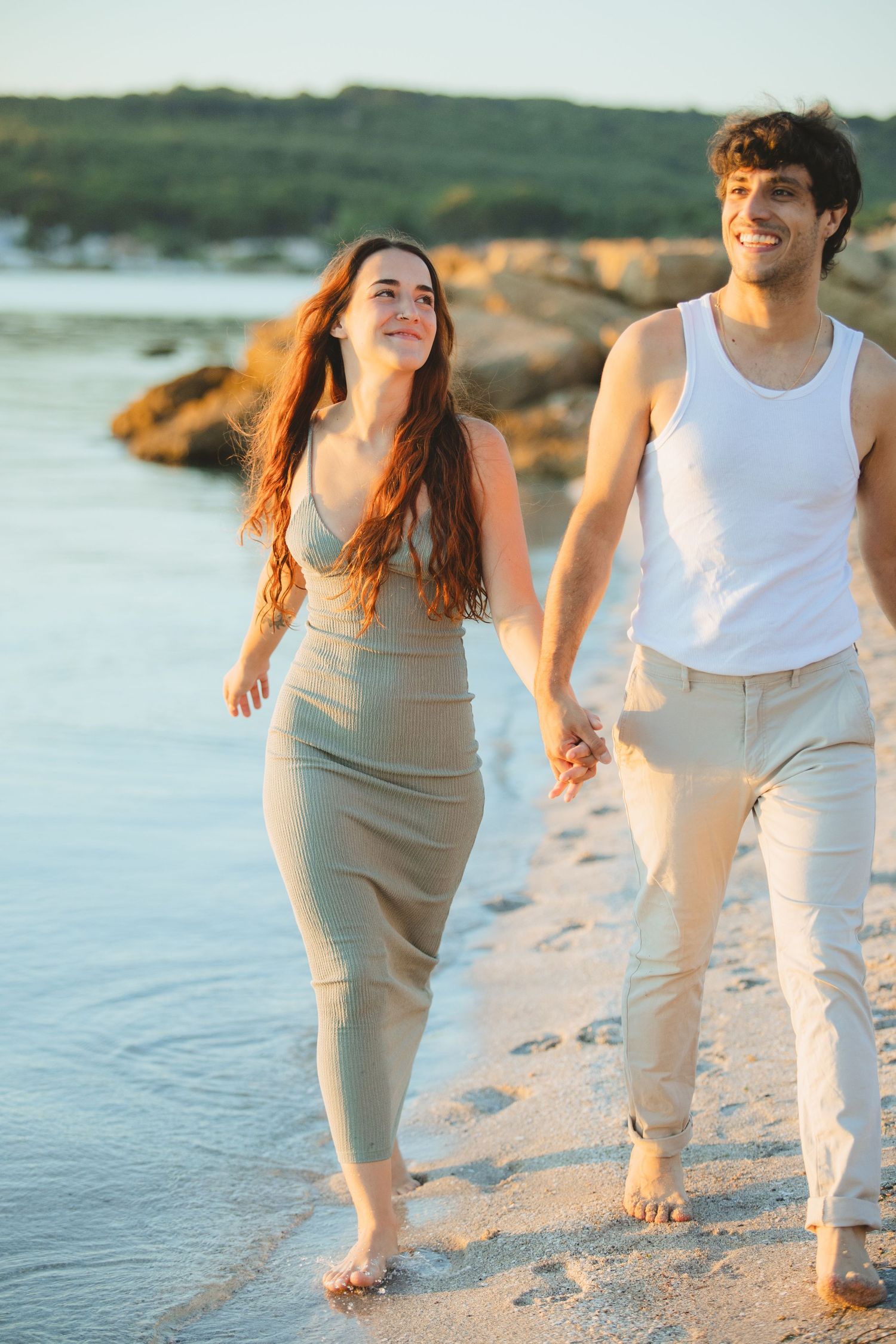 Happy couple holding hands walking barefoot on a sandy beach at golden hour sunset.