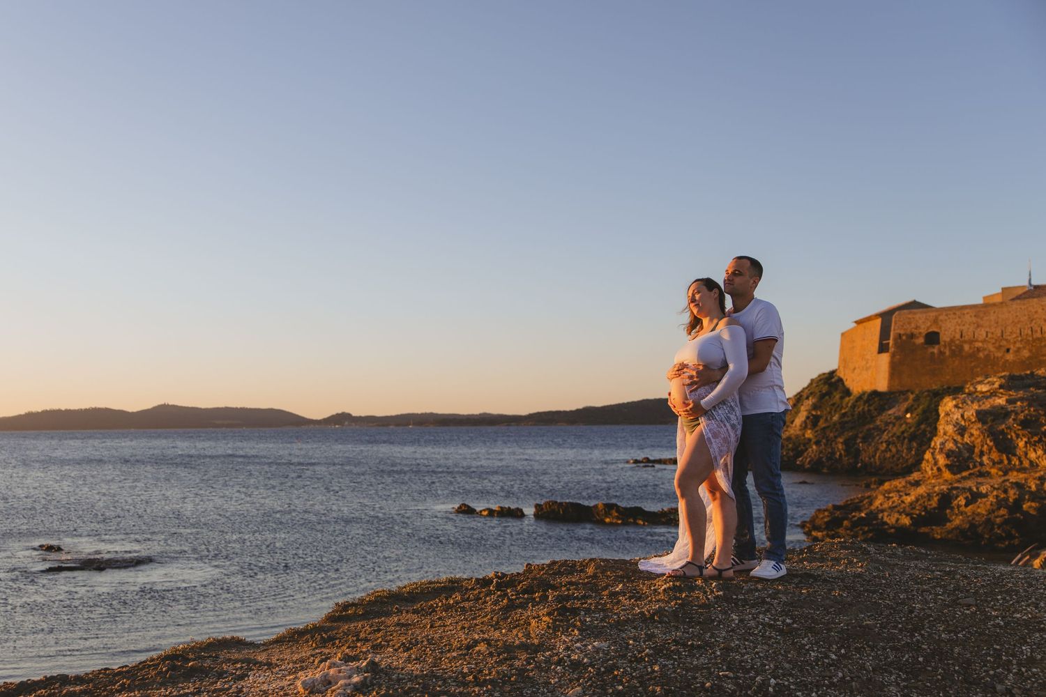 Couple standing together on rocky coastal shore admiring scenic sunset over the sea.