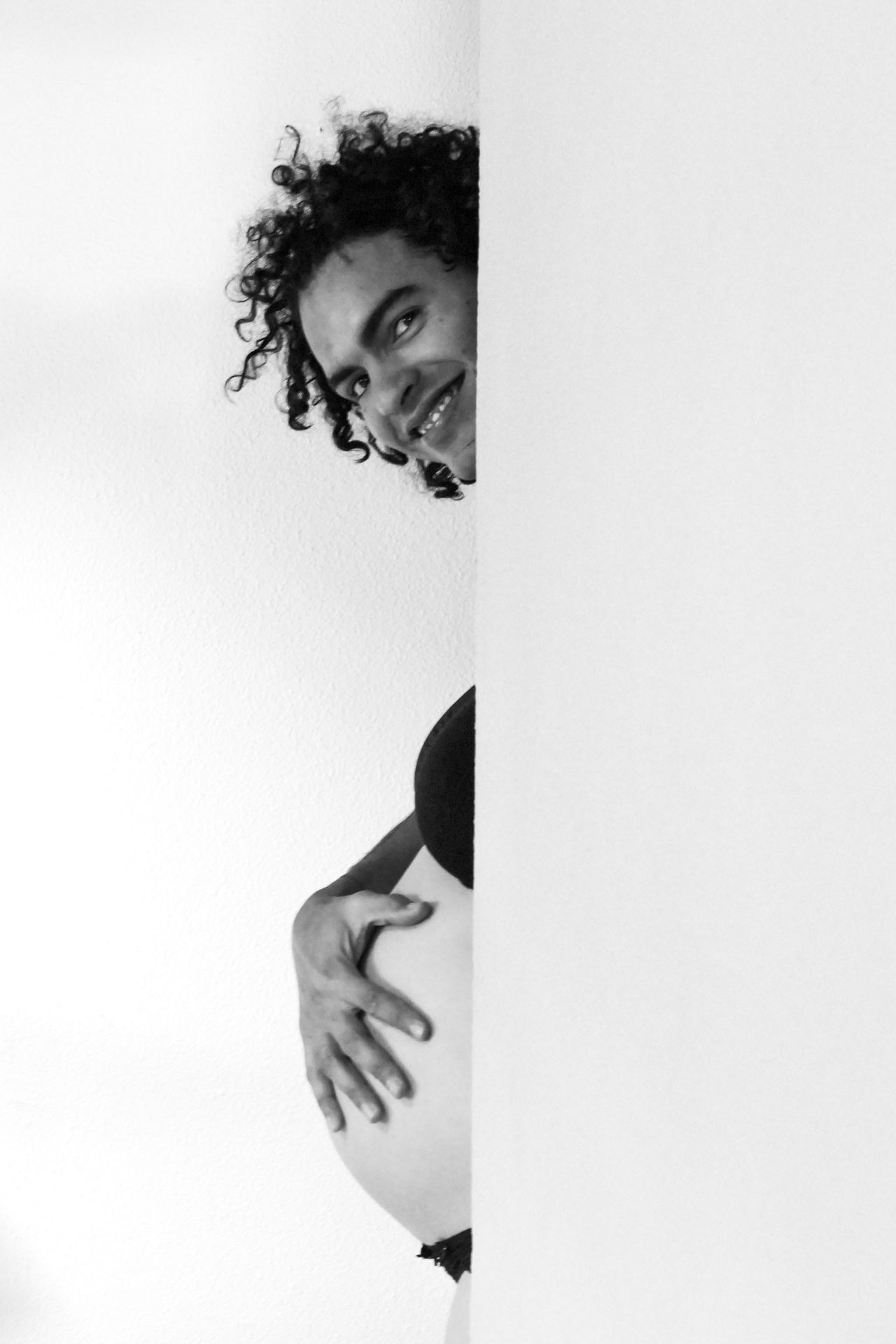 Smiling curly-haired pregnant woman peeking around white wall in black and white photo.