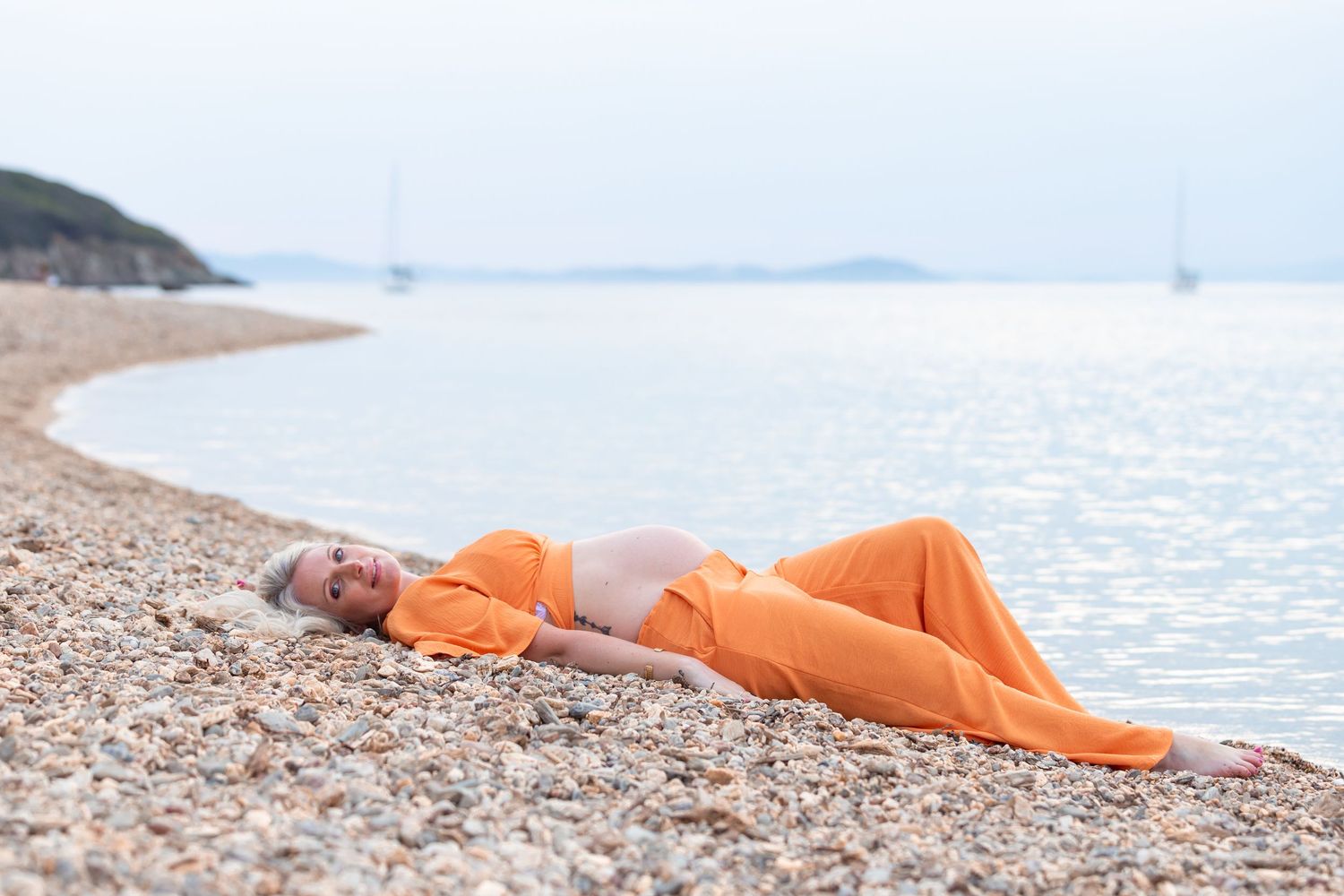 Pregnant woman in orange outfit lying on a pebble beach by calm sea with sailboats and green hills.