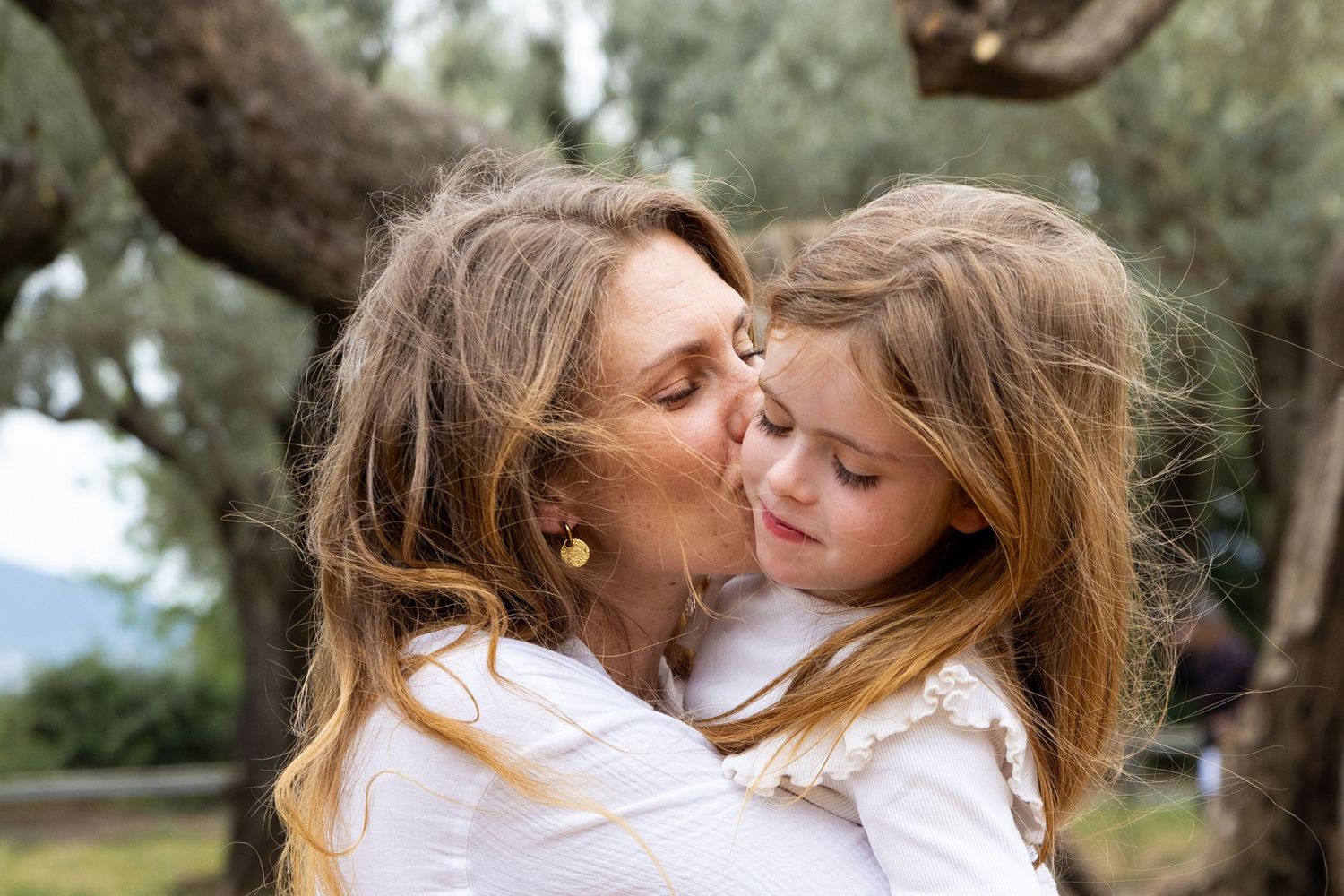 A mother tenderly kisses her young blonde daughter on the cheek outdoors among olive trees.