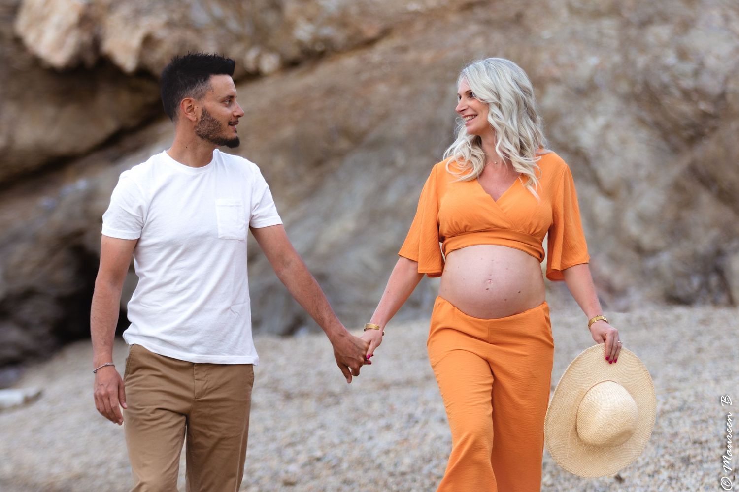 Happy expecting couple walking hand in hand on beach, woman in orange maternity outfit holding hat.