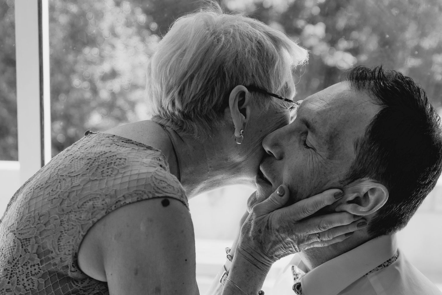 Elderly woman tenderly holding a man's face for a kiss in a black and white intimate portrait photo.